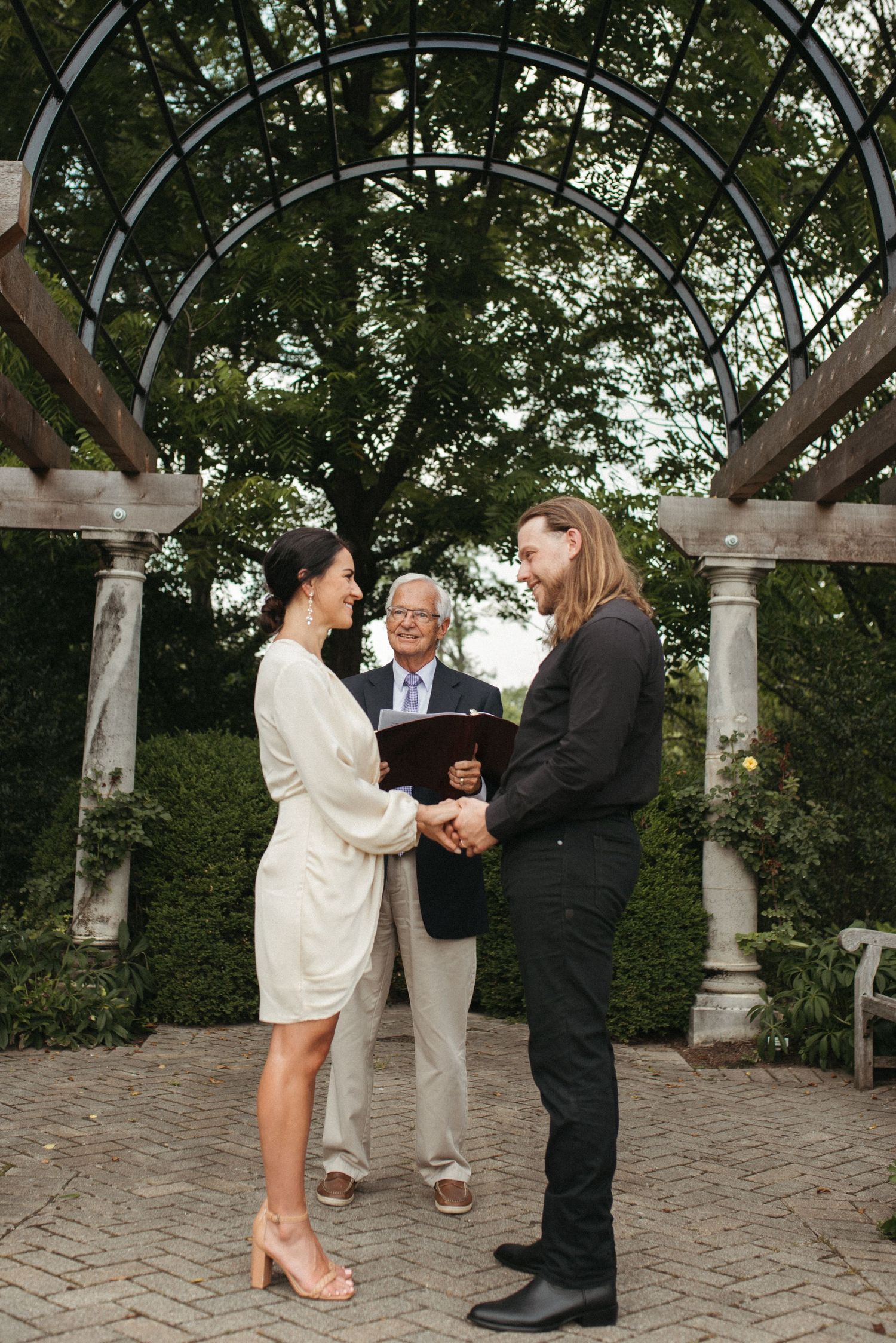 A wedding ceremony moment captured beneath an elegant garden pergola with curved metal beams.