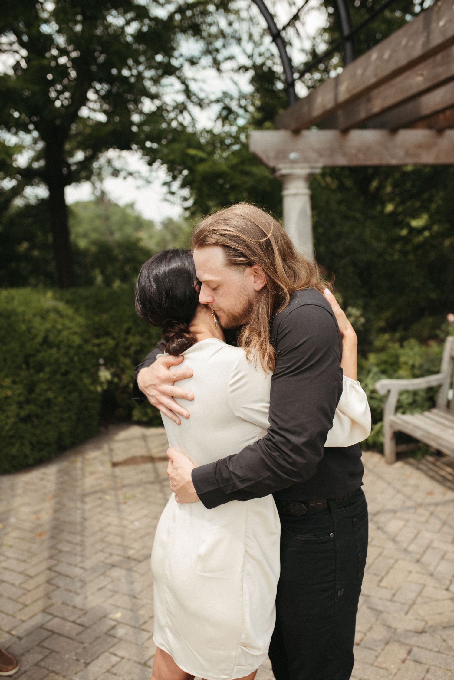 An intimate embrace between a couple in a garden with stone columns and greenery.