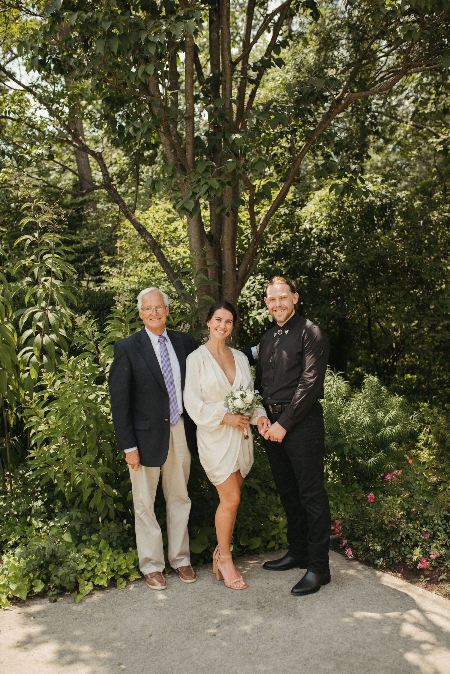 A wedding party of three poses outdoors in formal attire with lush greenery and trees in the background.