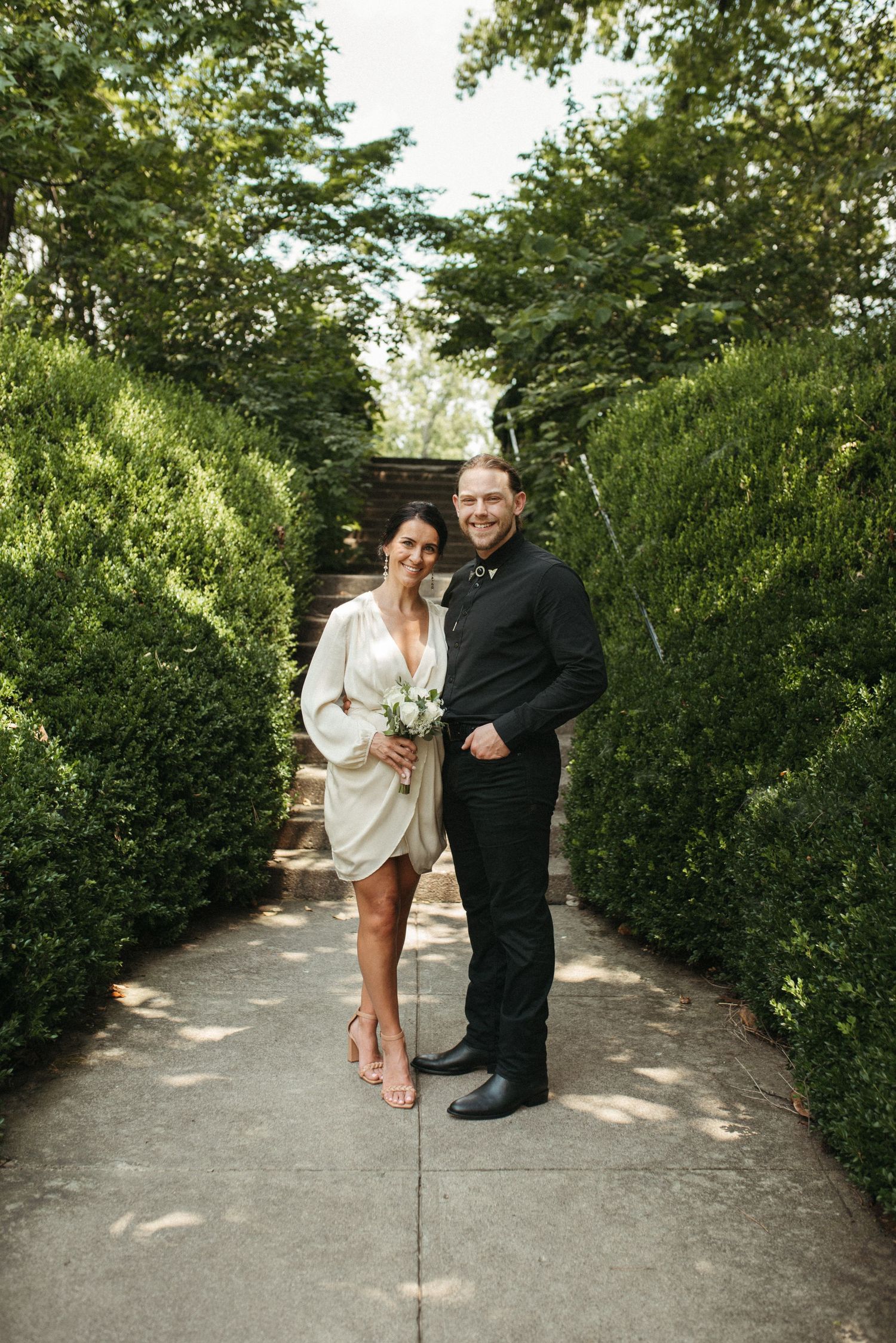 A couple poses on a garden path lined with manicured hedges and stairs in the background.