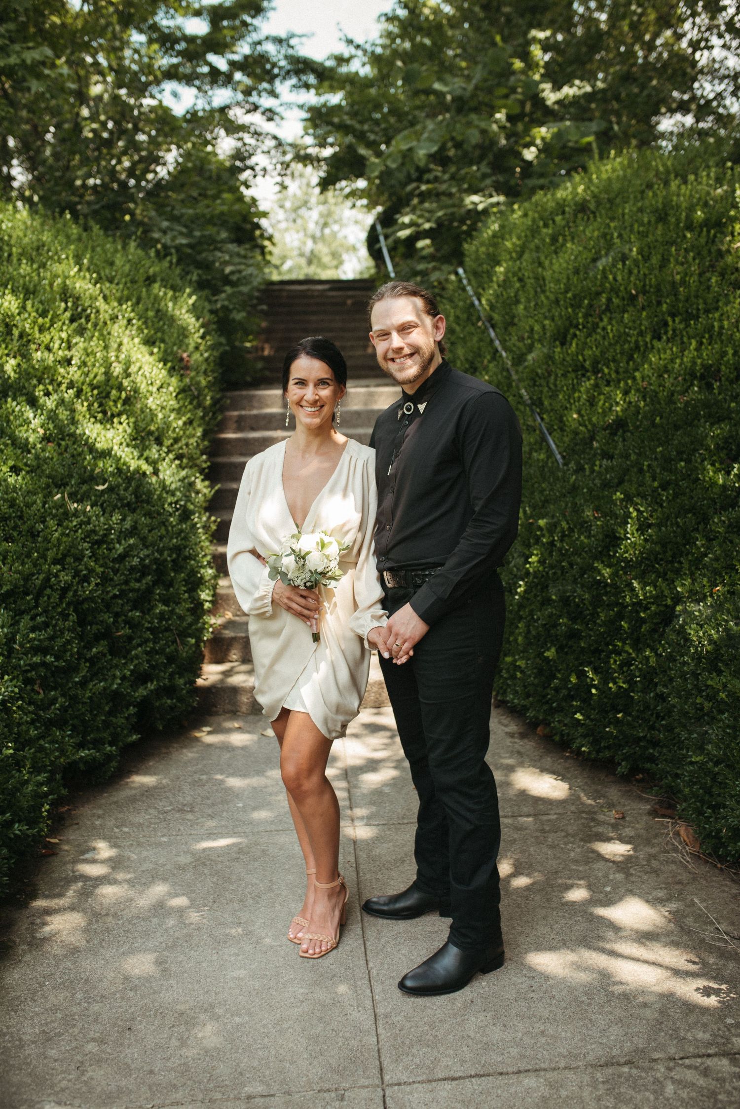 A couple stands together on a stone pathway surrounded by green hedges and garden steps.