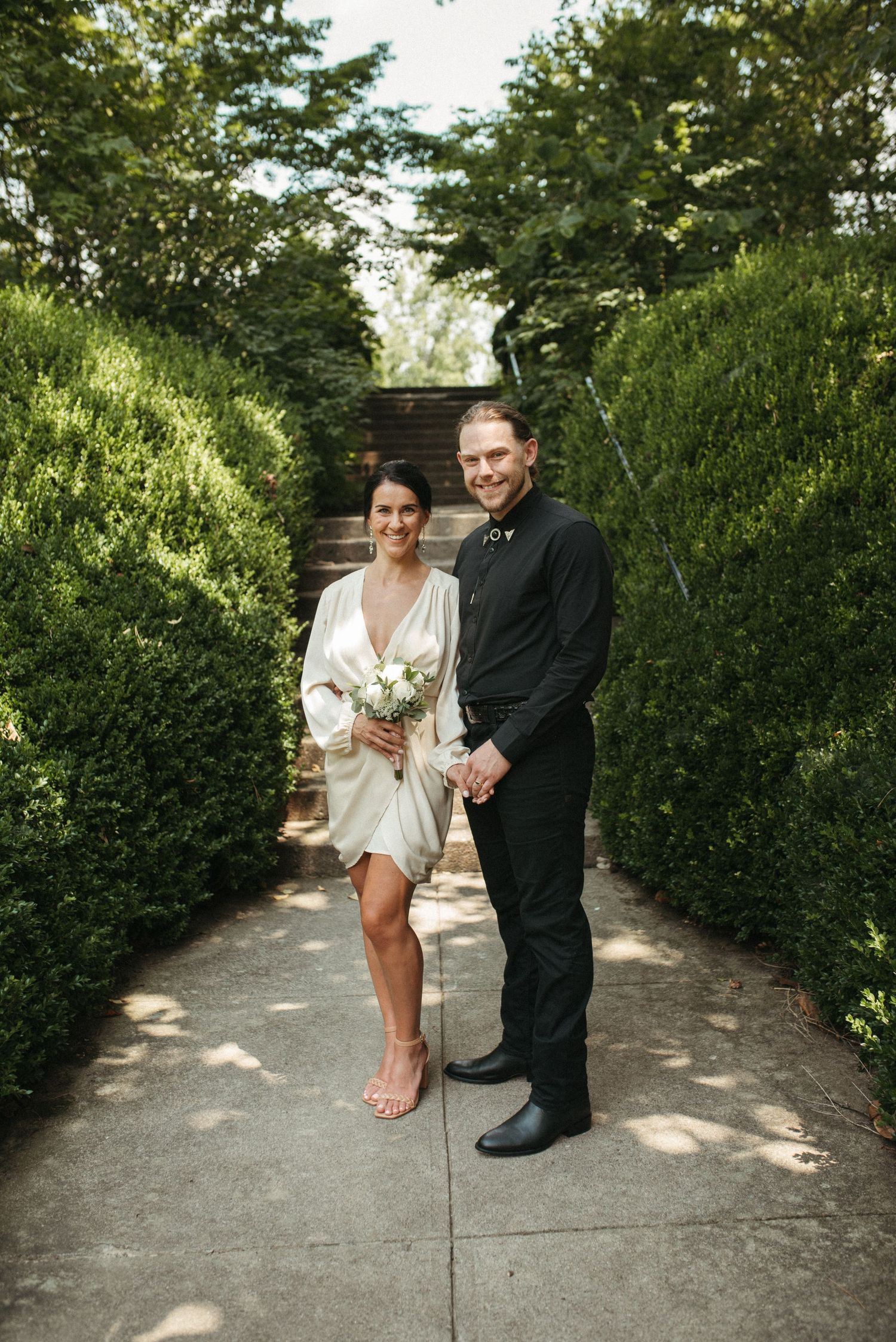 A couple poses together on a garden path with stone steps and hedges creating a natural backdrop.