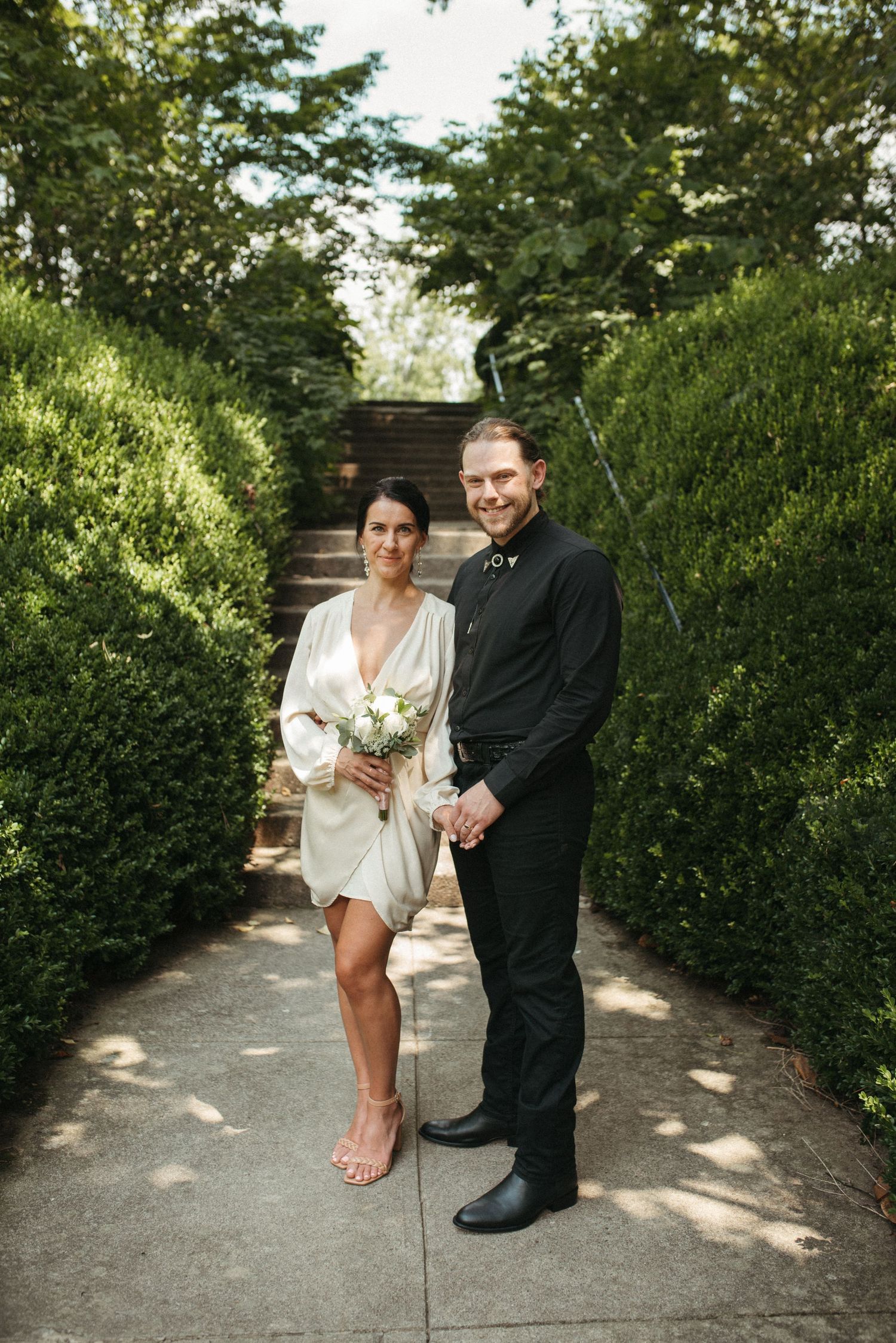 A couple stands smiling on a garden pathway with stone steps and manicured hedges.