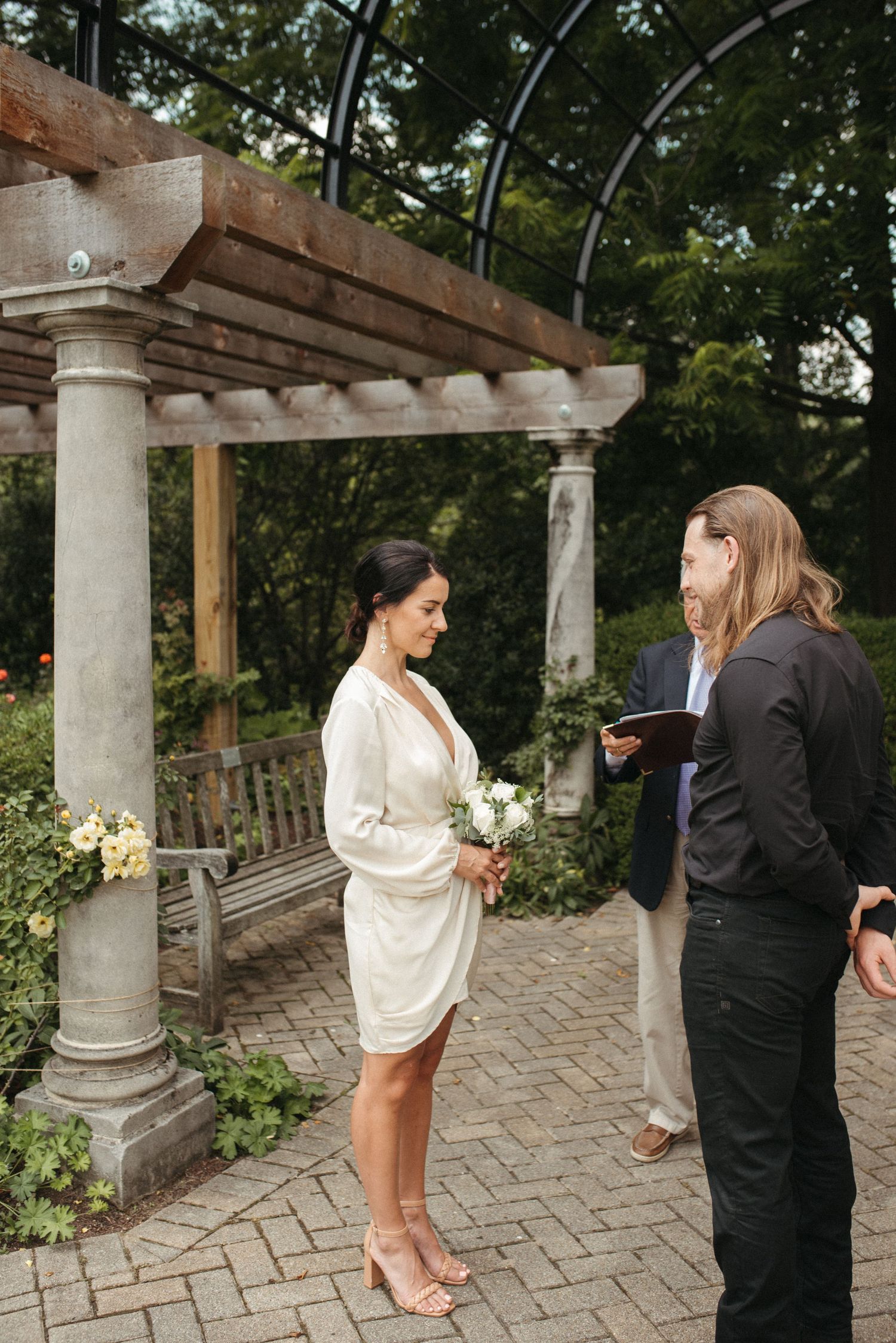 An intimate wedding ceremony unfolds beneath a rustic garden pergola with stone columns.