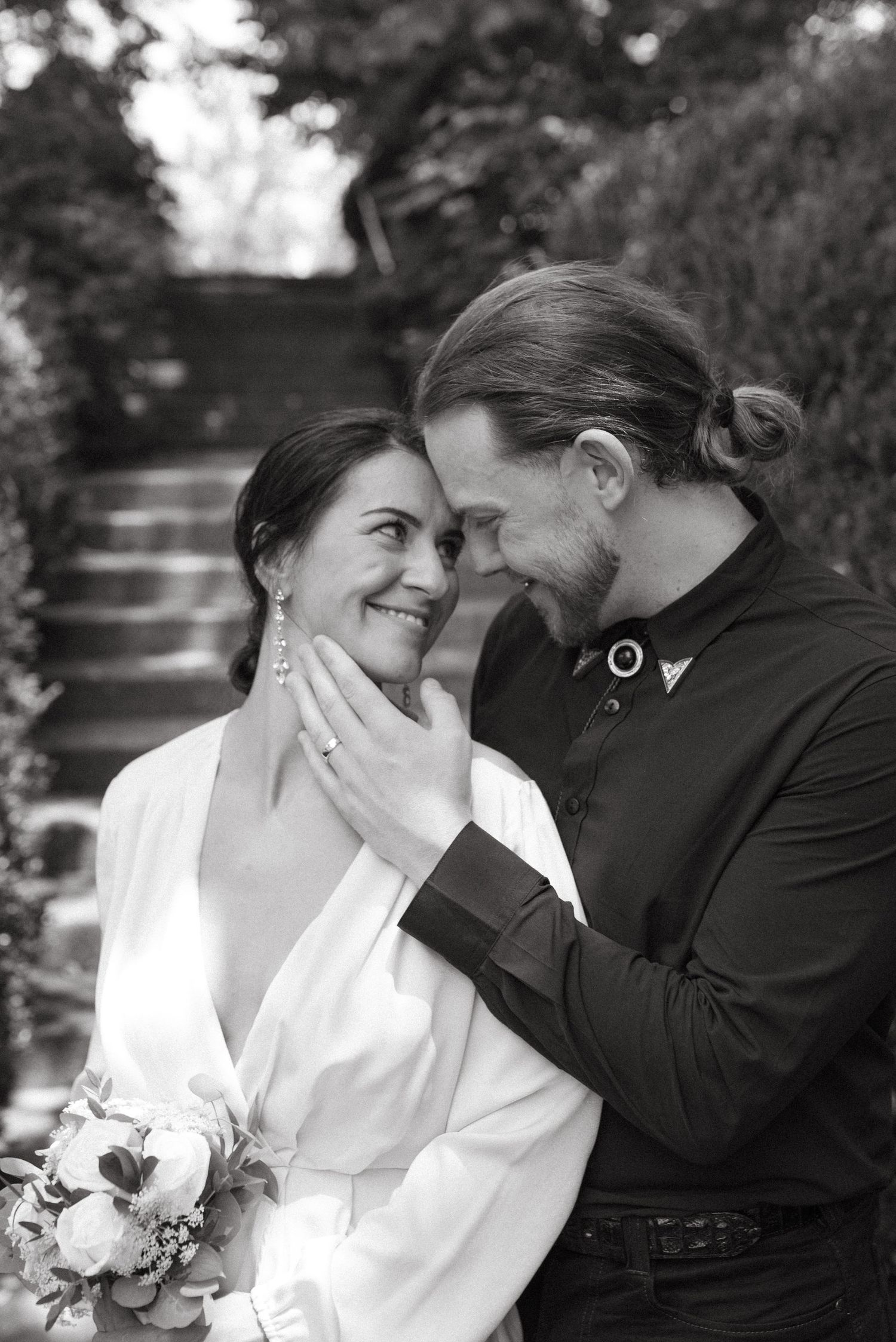 A black and white portrait of a couple in formal attire sharing a tender moment outdoors.