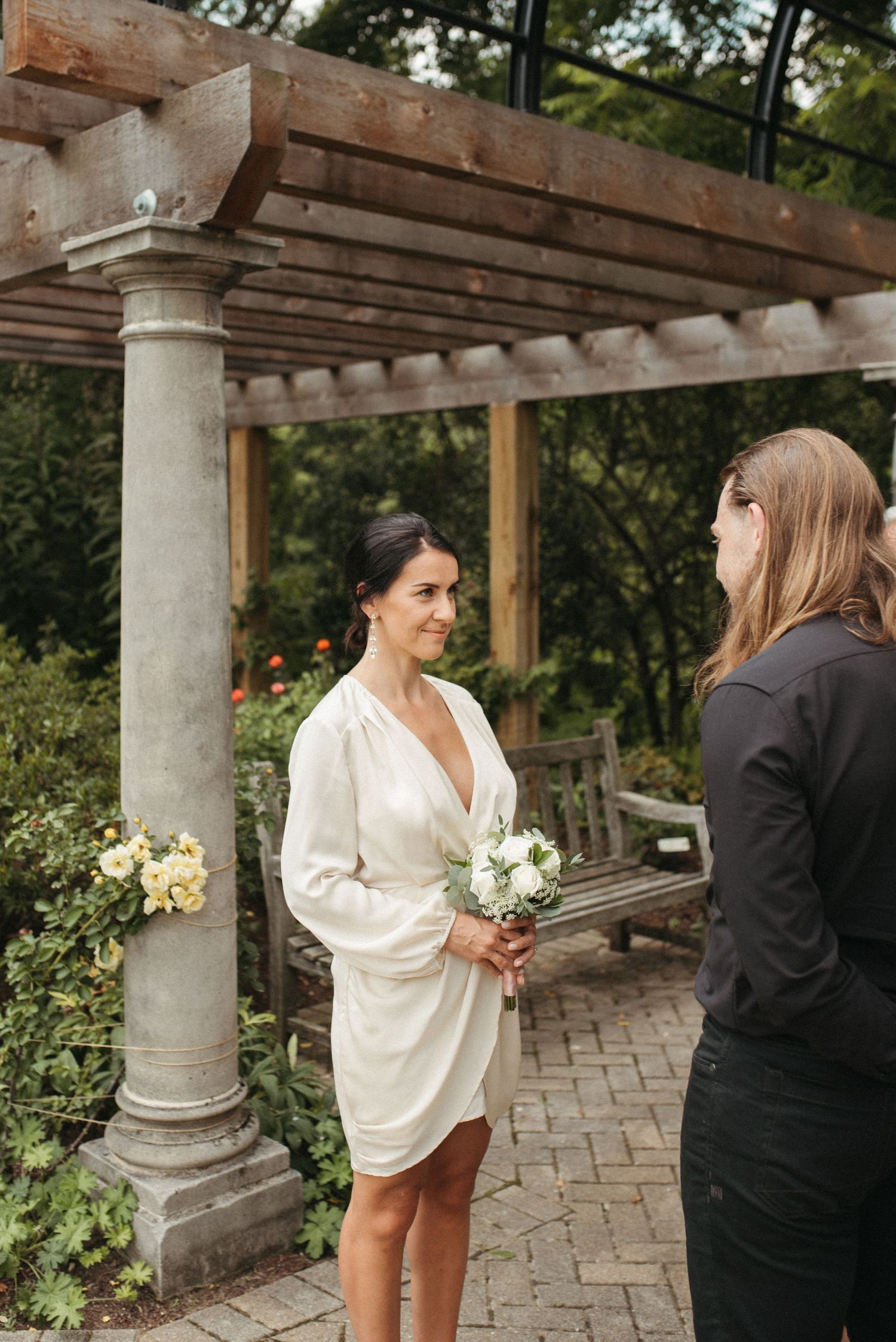 A wedding ceremony takes place under a stone pergola with wooden beams in a garden setting.