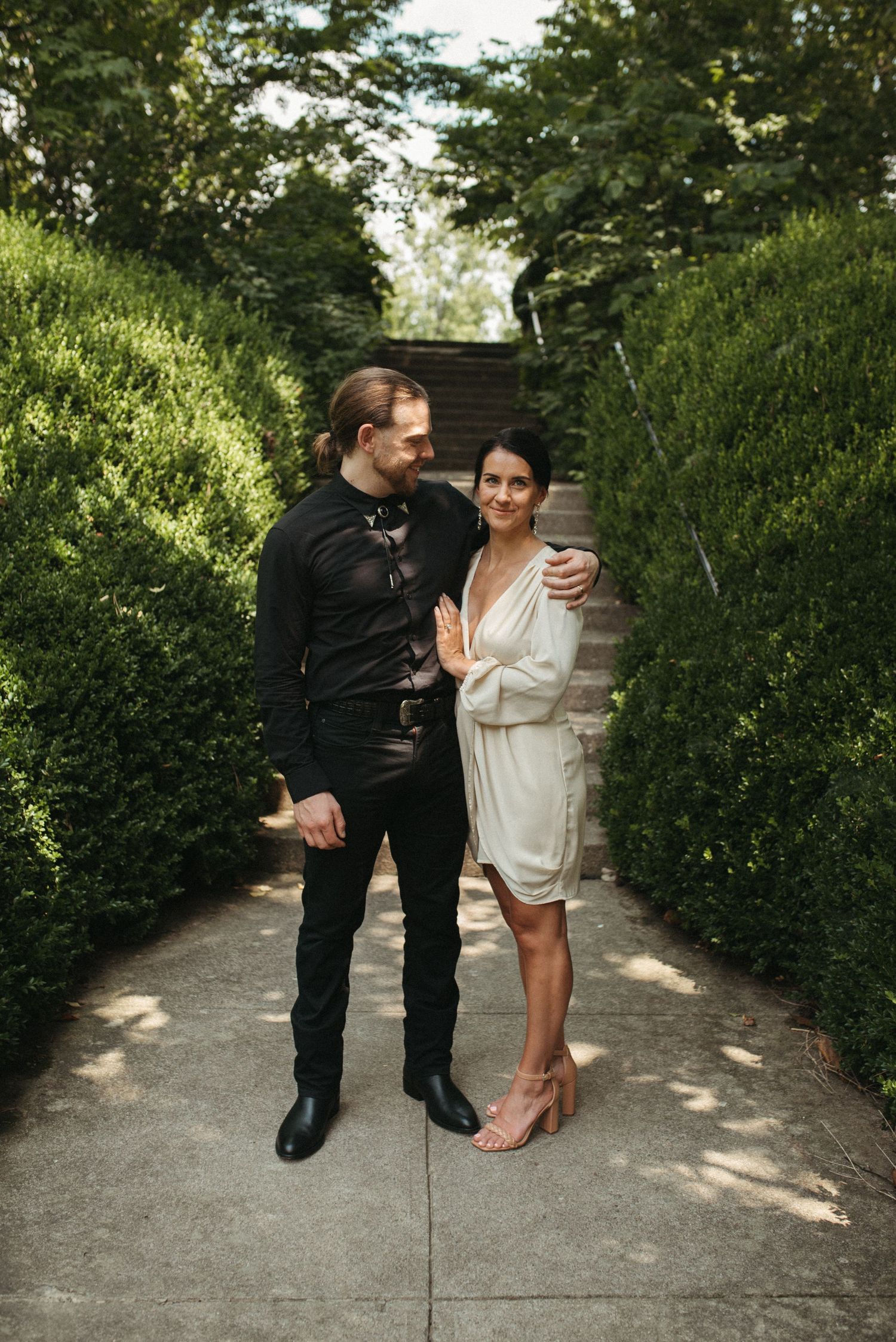 A full-length portrait of a wedding couple standing on a garden path lined with hedges.
