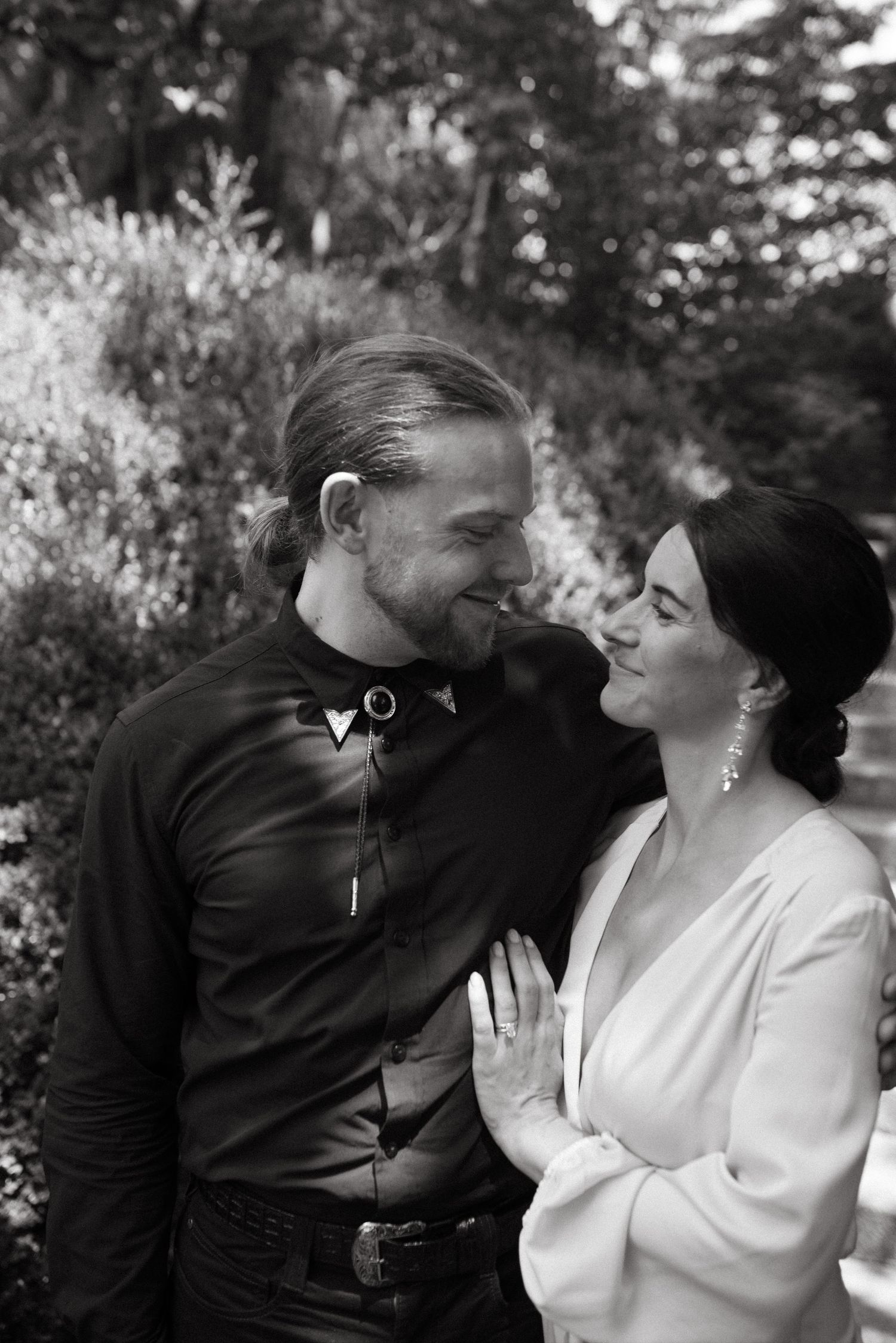 A black and white portrait of a romantic couple embracing outdoors in a garden setting.