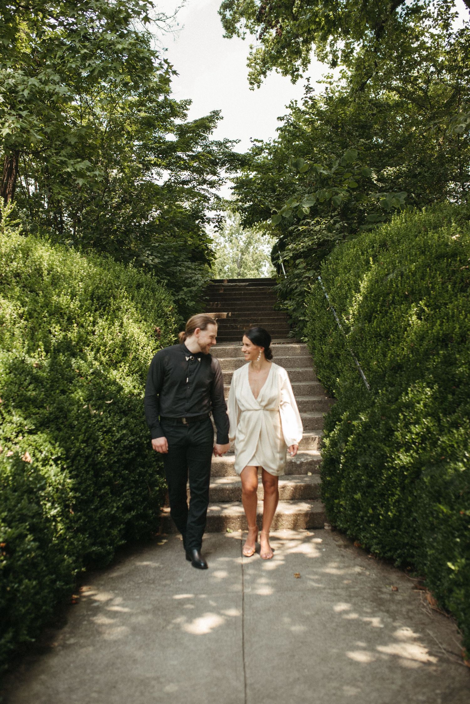 A couple walks hand in hand down stone garden steps surrounded by lush greenery.