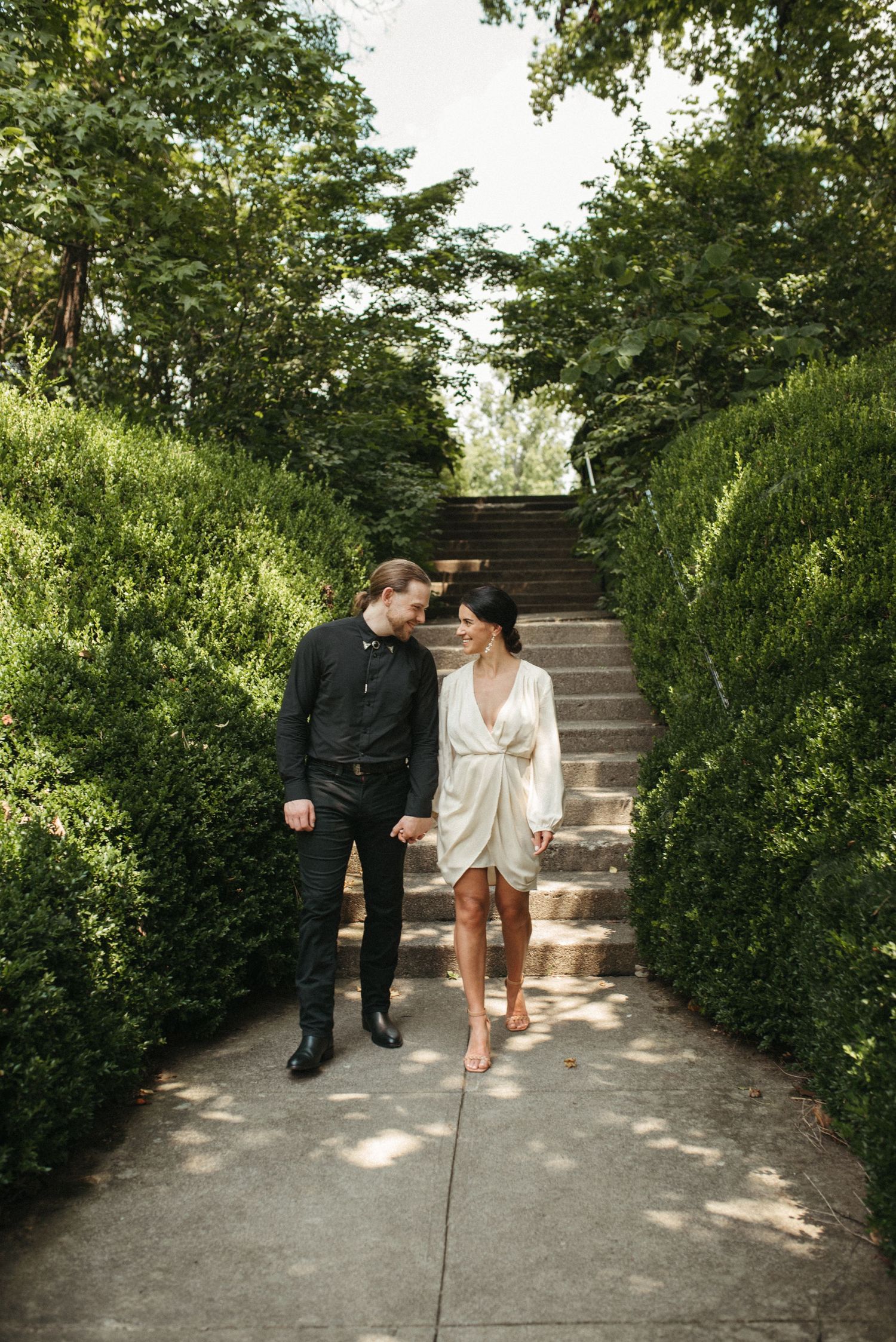 A couple in contrasting outfits descends garden stairs while sharing an intimate moment.