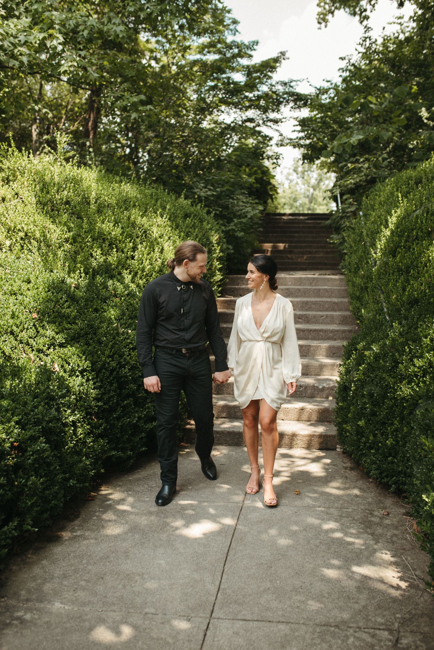 A stylish couple walks together on a garden path with stone steps in the background.