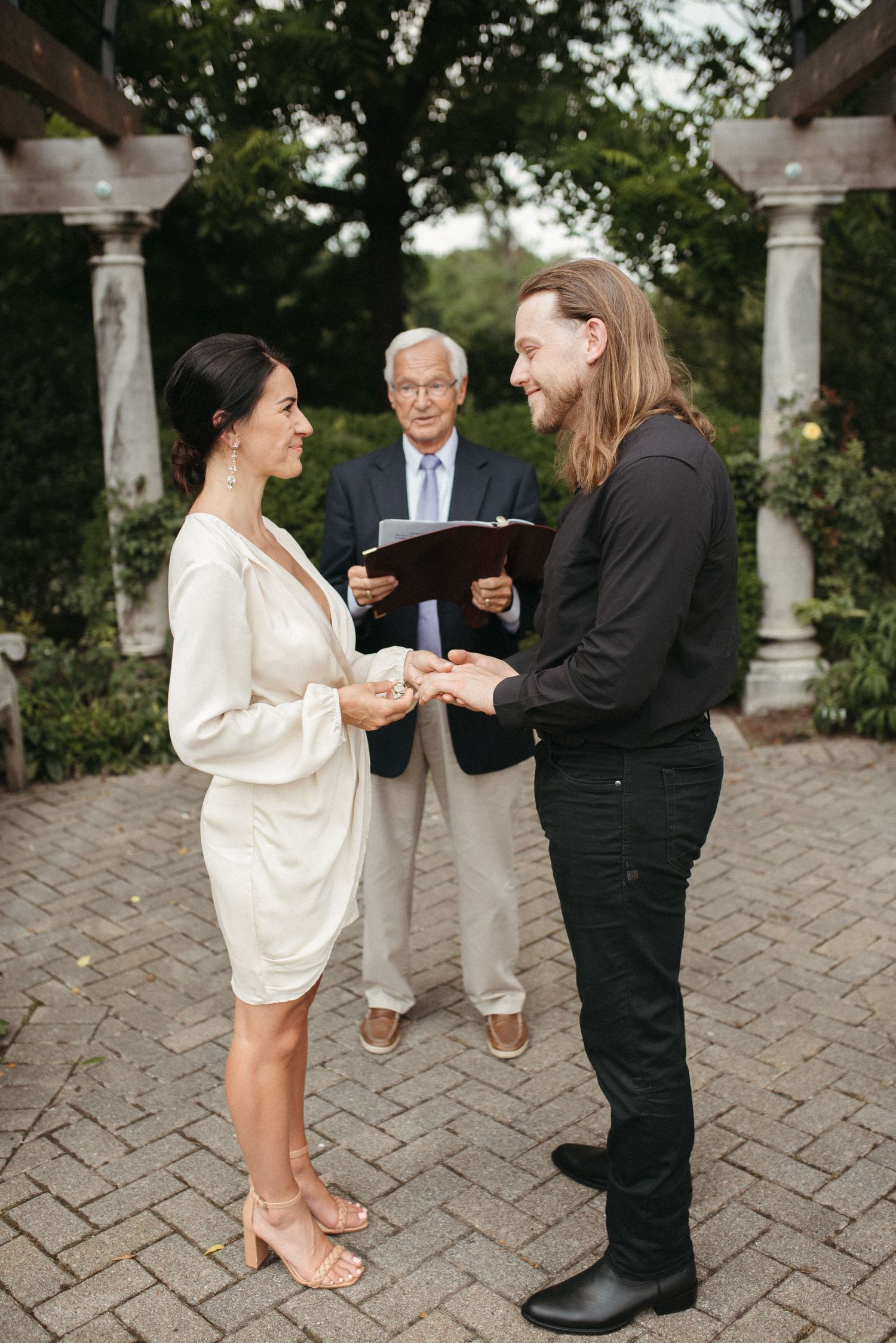 A small wedding ceremony takes place under an elegant pergola with stone pillars and wooden beams.