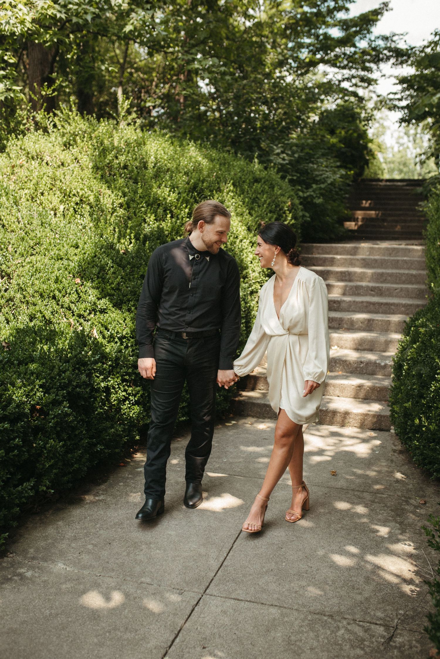 A couple in elegant attire strolls together along a garden pathway with stone steps.
