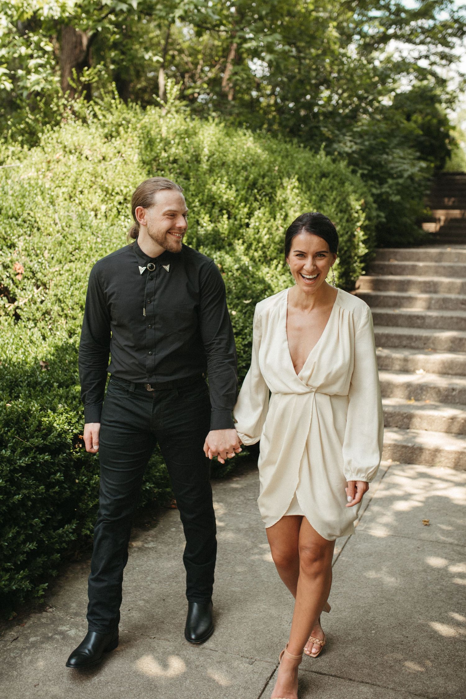 A happy couple holds hands while walking along a sunlit garden path with stone steps.
