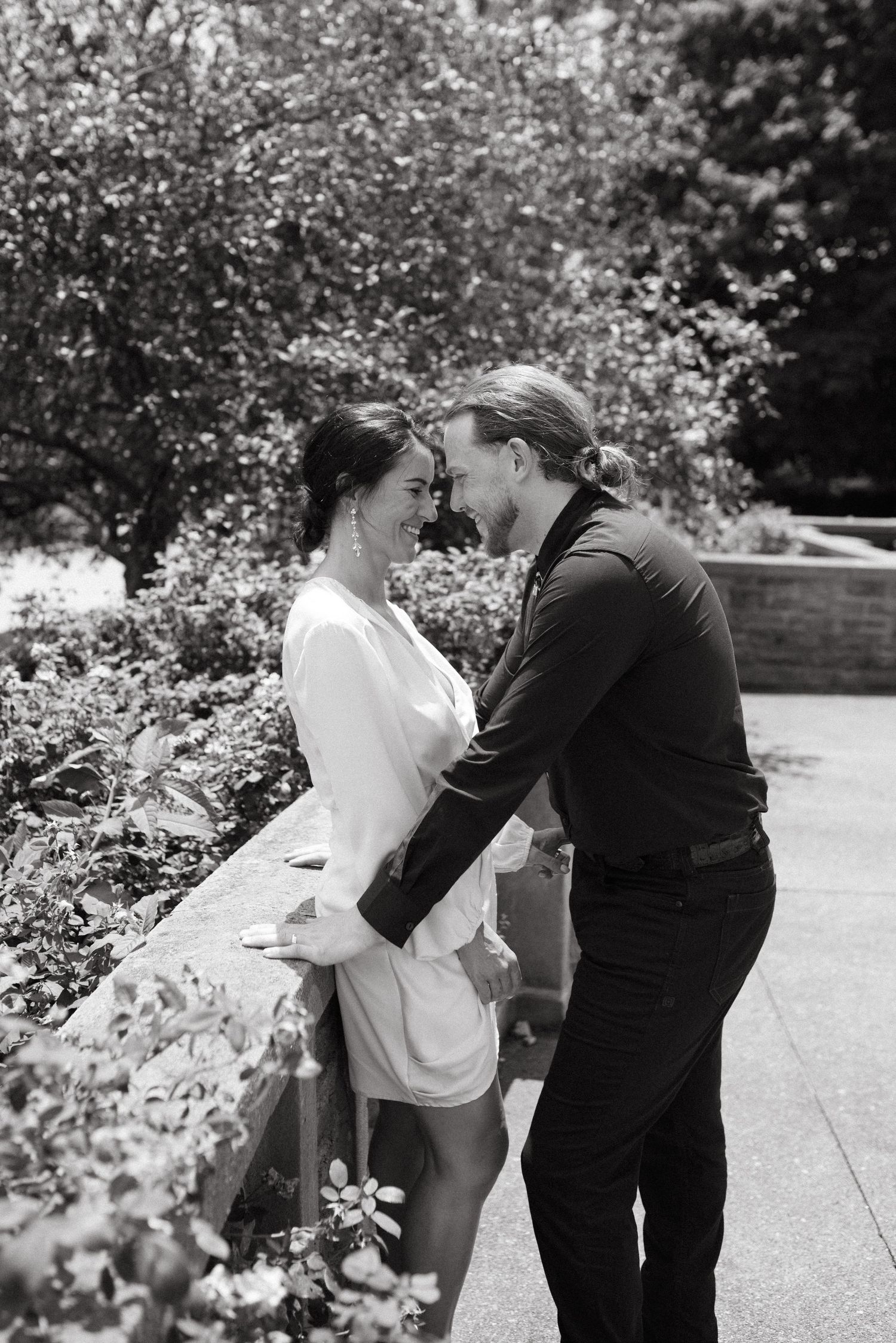 Black and white photo of a couple sharing an intimate moment by a garden wall.