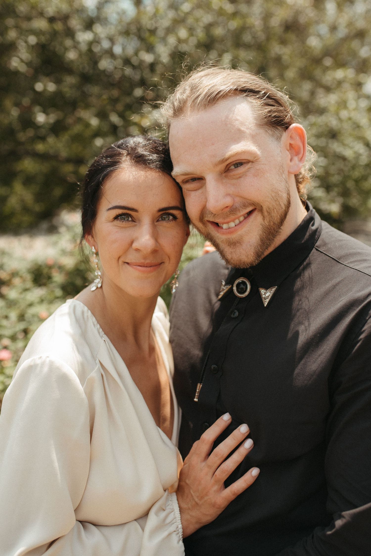A couple dressed in black and cream poses for a close-up portrait in a garden setting.