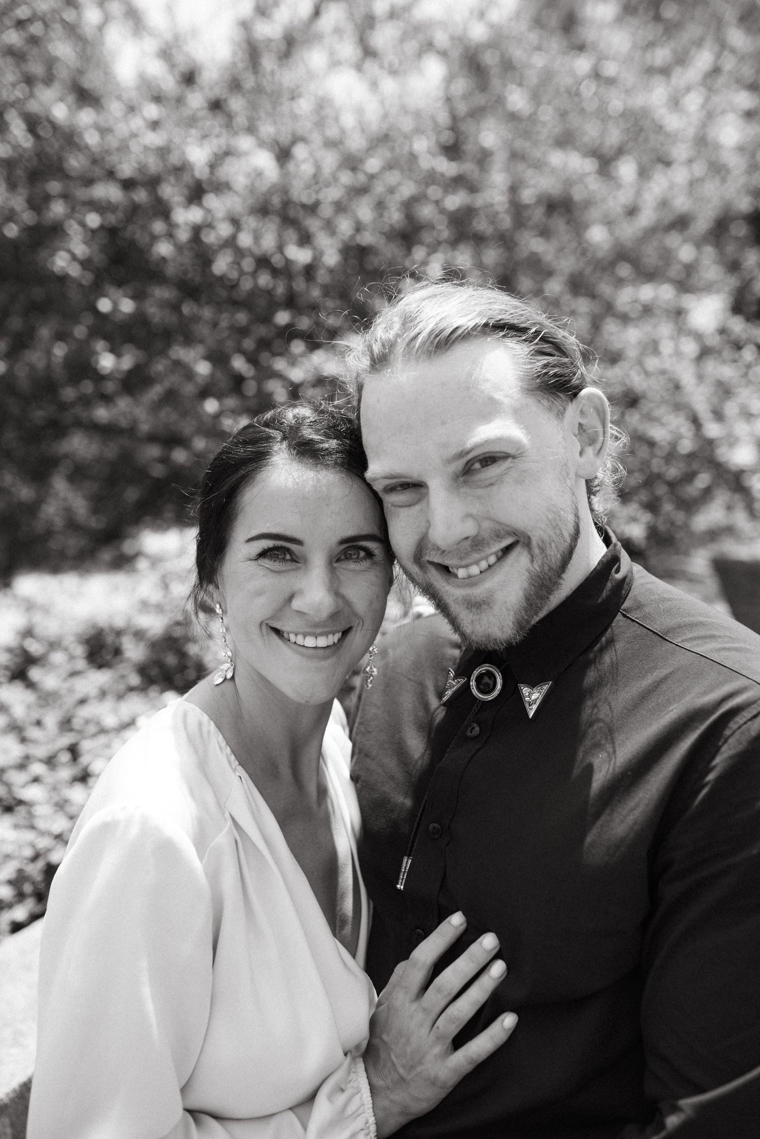 A romantic black and white portrait of a couple in formal attire smiling at the camera outdoors.