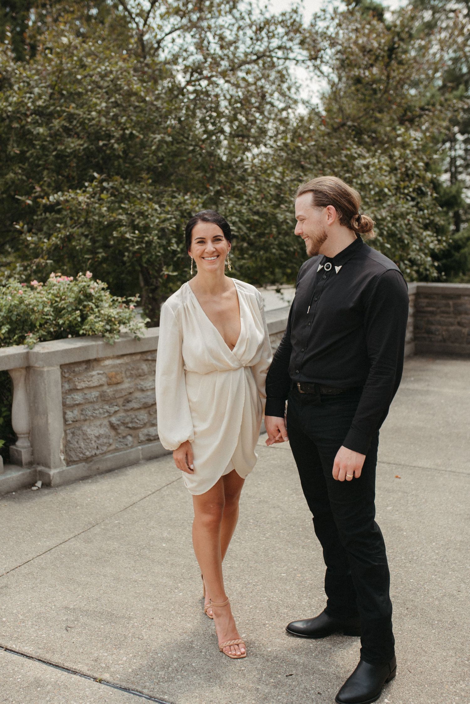 Couple in contrasting outfits walking together on a stone terrace surrounded by trees.