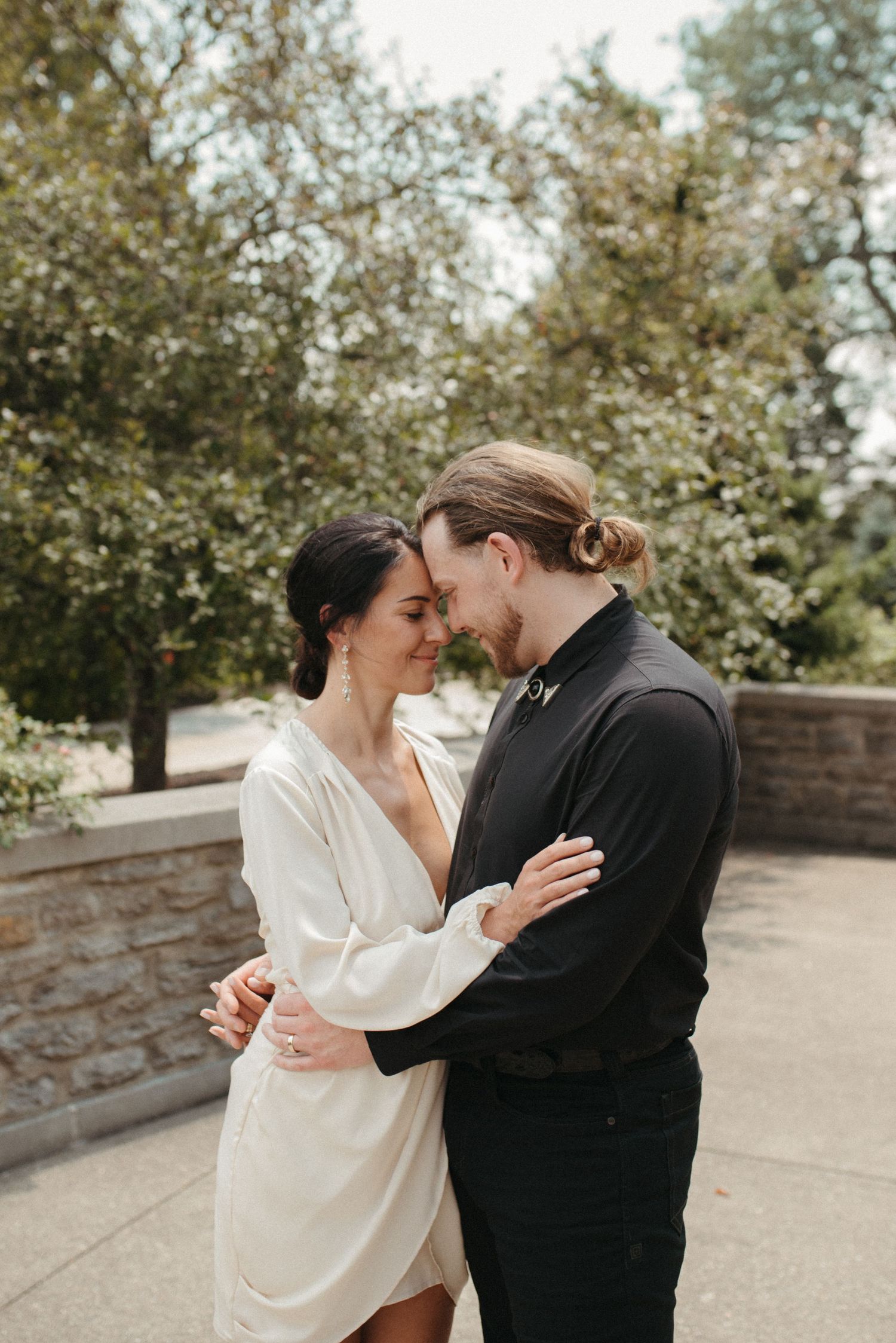 An intimate moment between a couple in black and white dress embracing near a stone wall and trees.