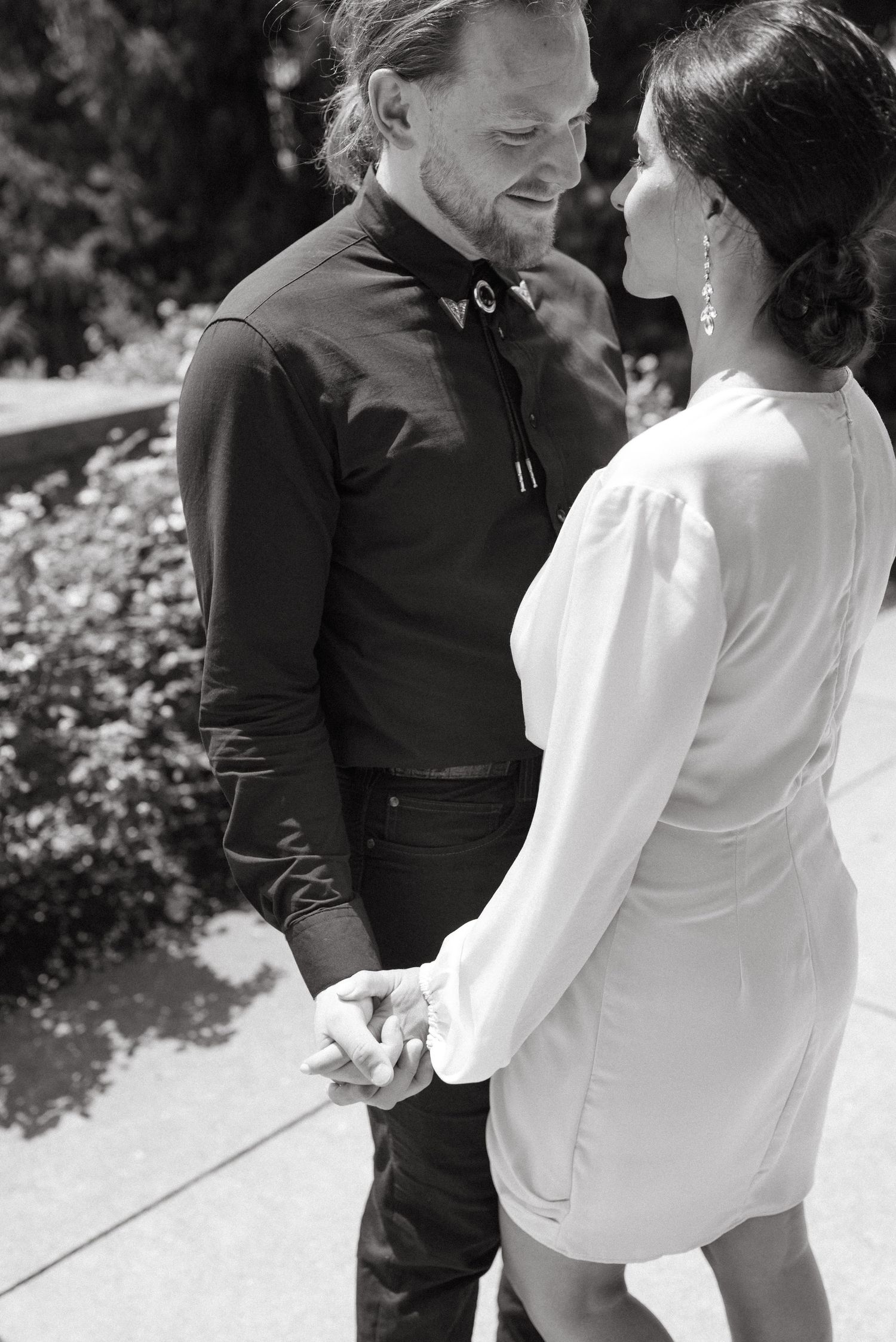 A tender black and white moment between a couple in formal wear holding hands on a sunny path.