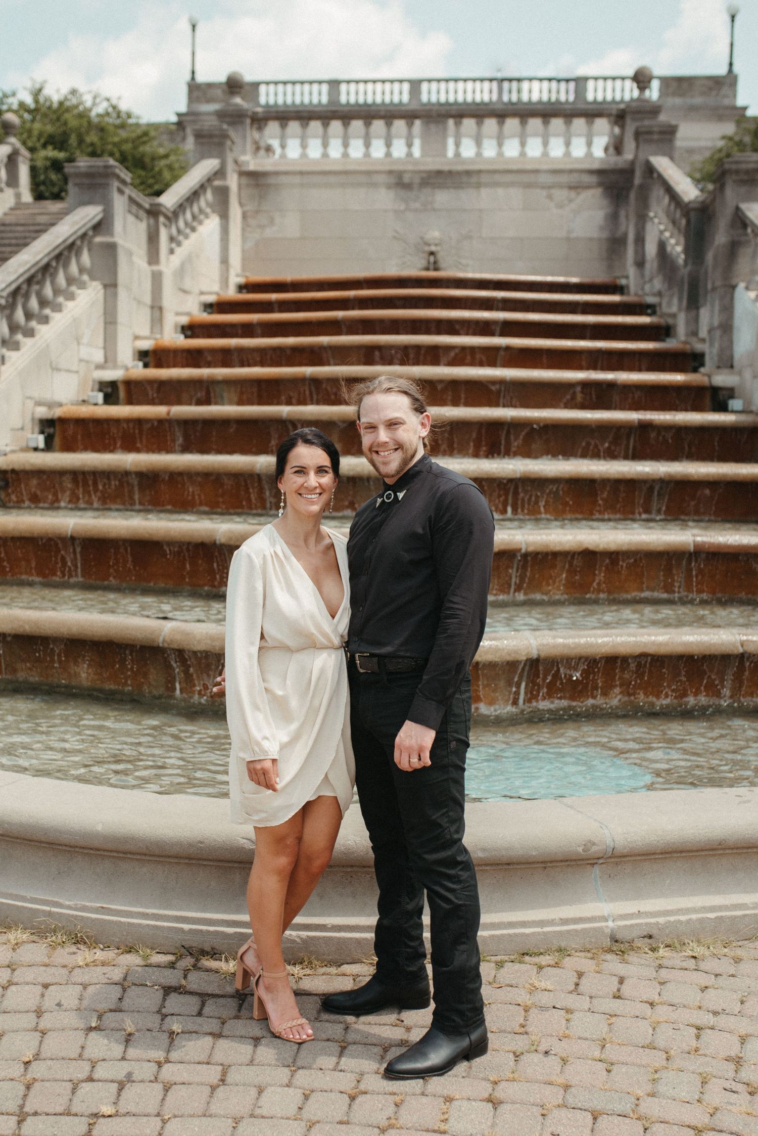 A couple poses together at the bottom of an ornate water fountain with cascading steps.