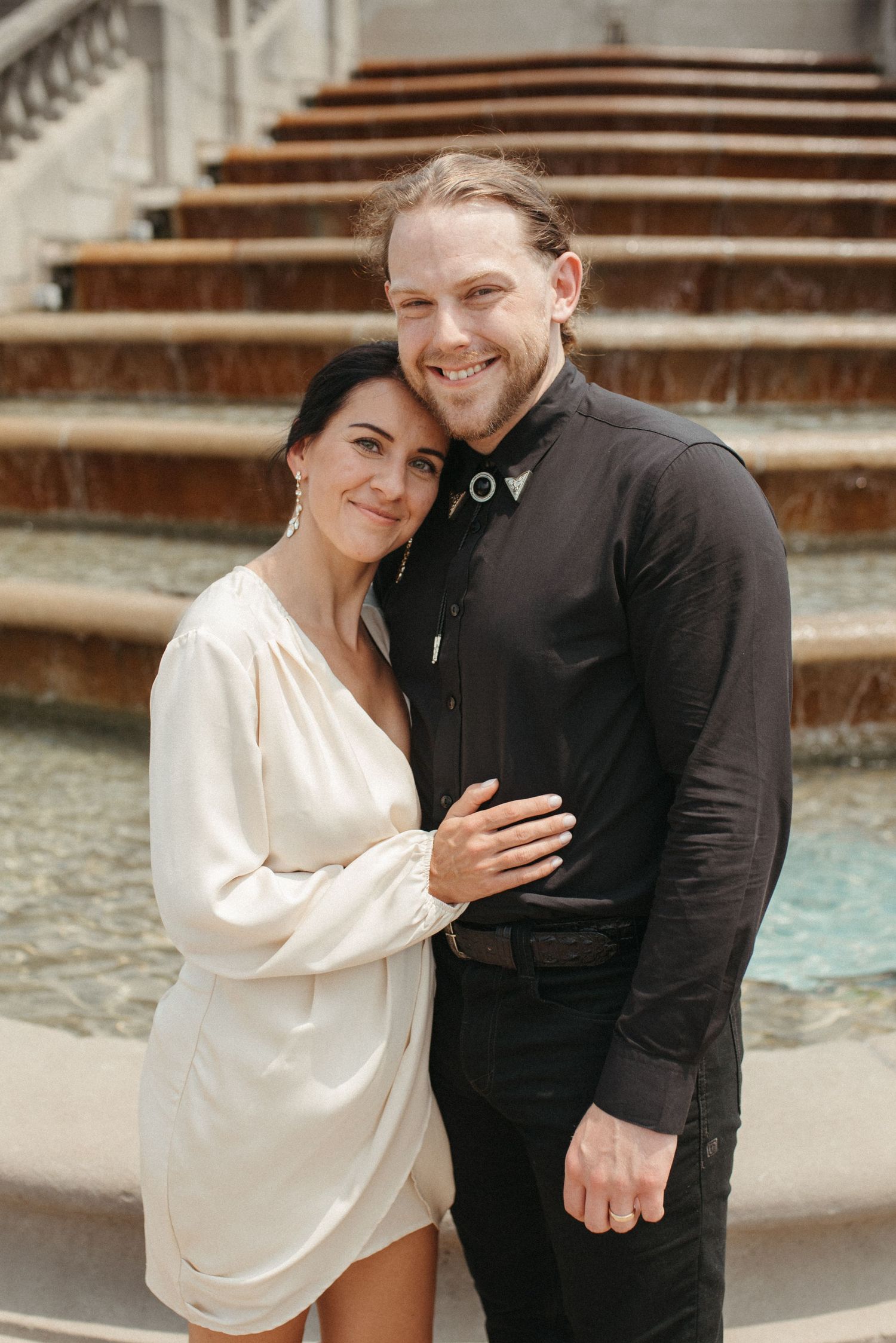 A happy couple embraces in front of a grand fountain with cascading steps.
