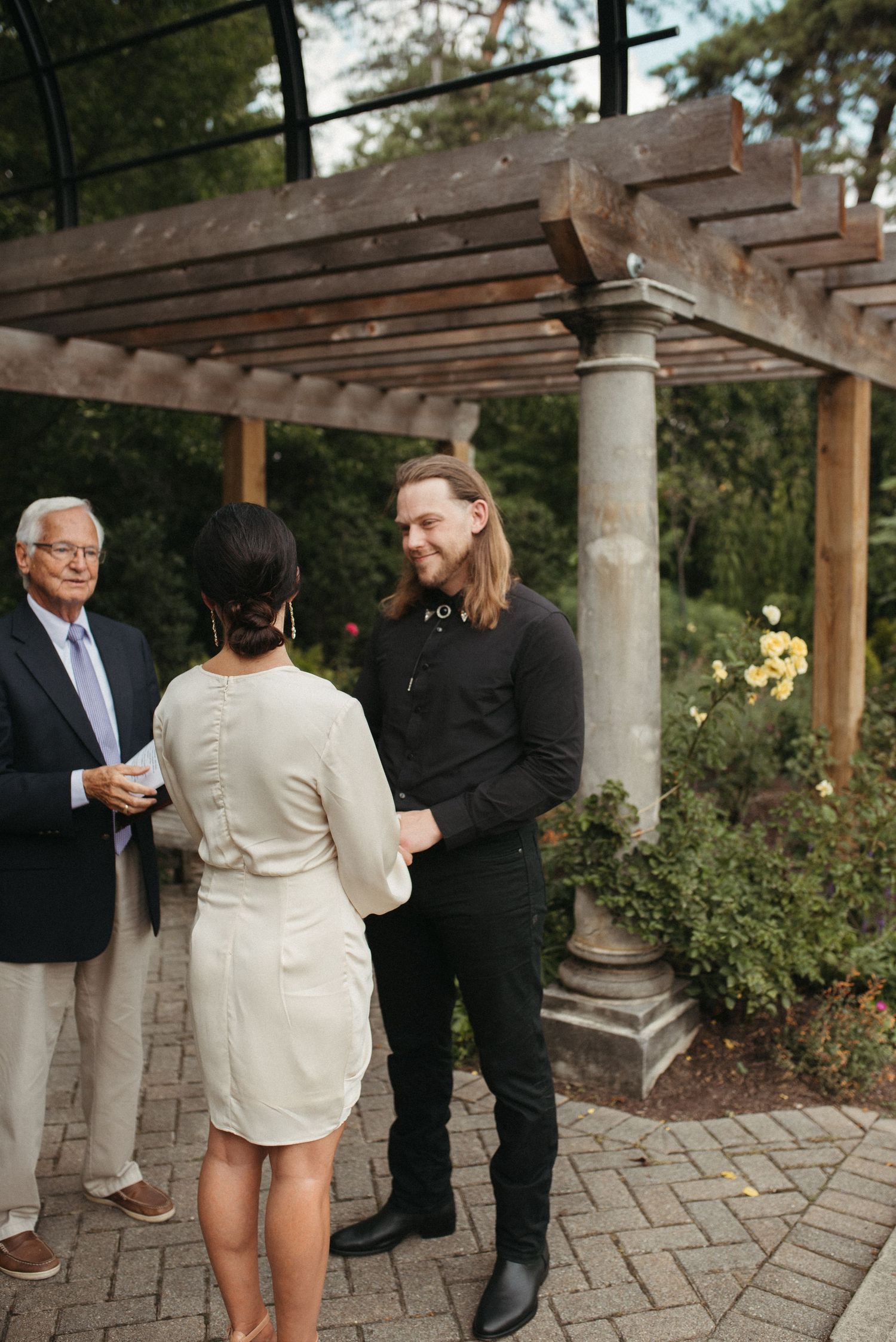 A wedding ceremony occurs under a wooden pergola with classical stone columns.