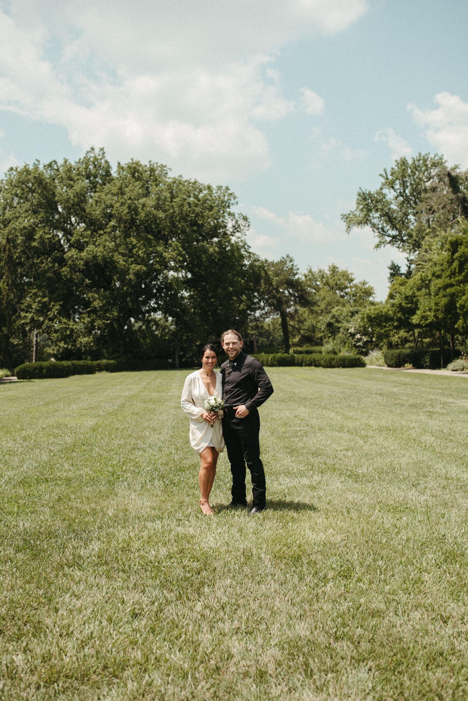 A couple poses together on a sunny day in a lush green park setting wearing casual formal attire.