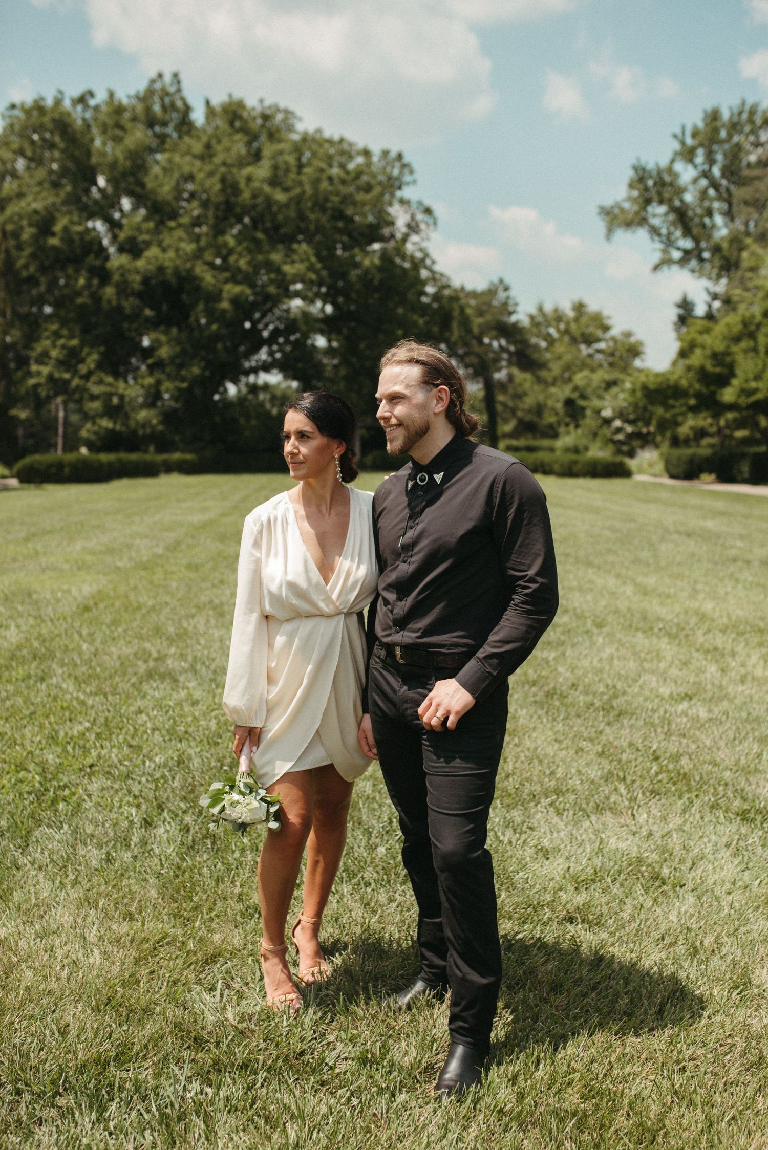 A couple in contrasting black and white outfits stands together on manicured grass under blue skies.