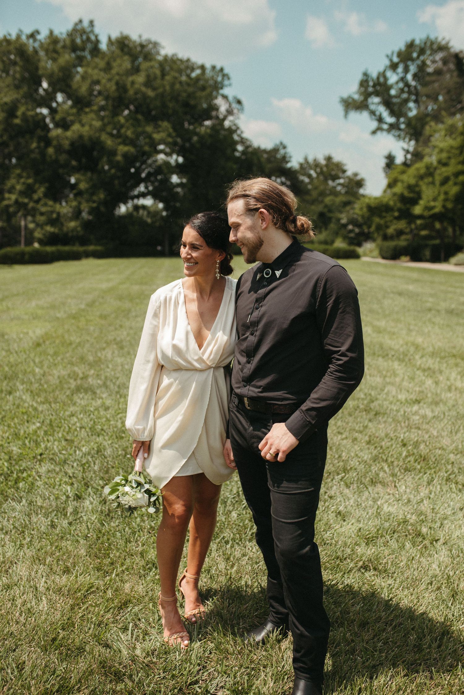 A couple walks hand in hand across a sunlit lawn surrounded by trees.