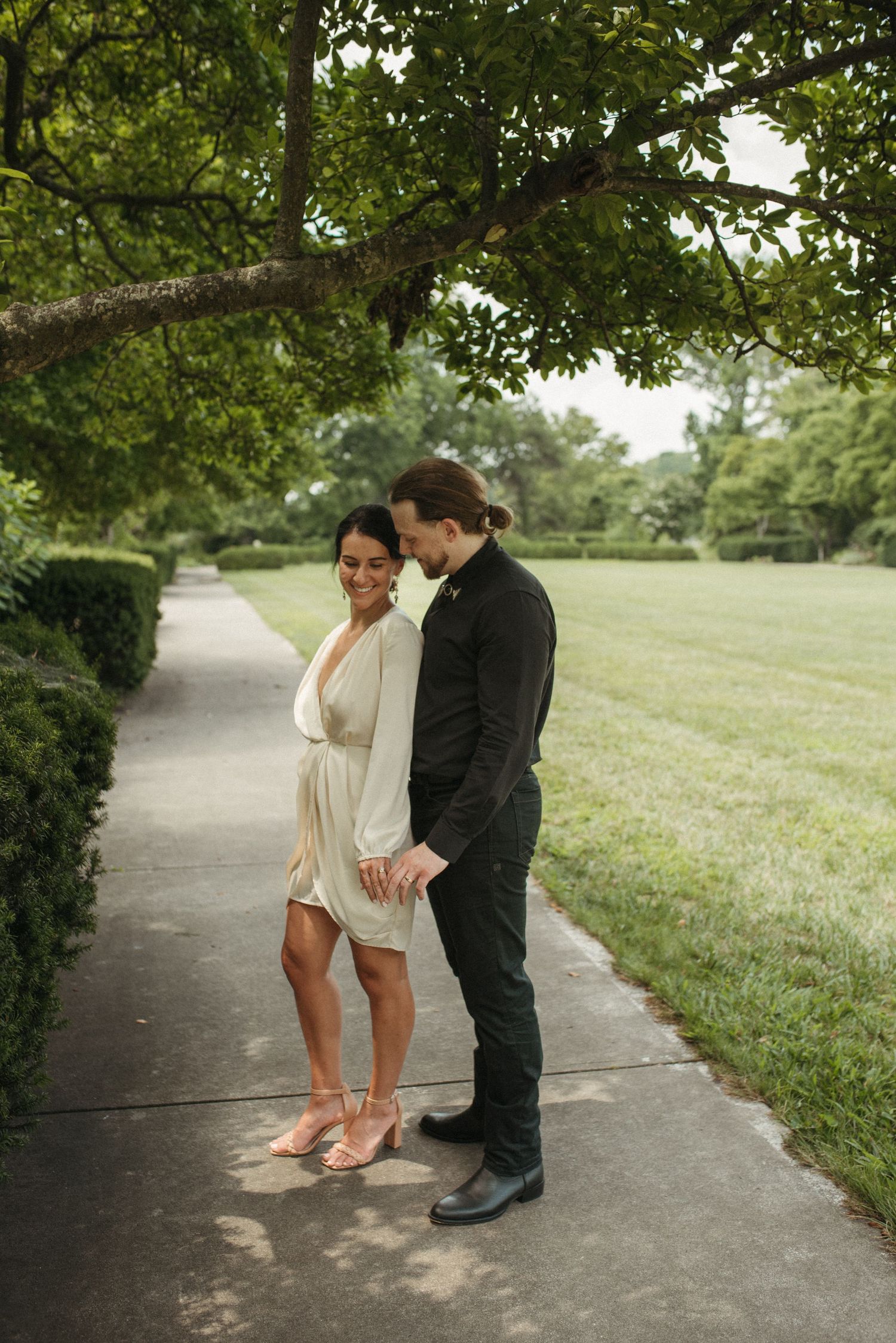 A couple stands together on a concrete path bordered by green lawn and trees.