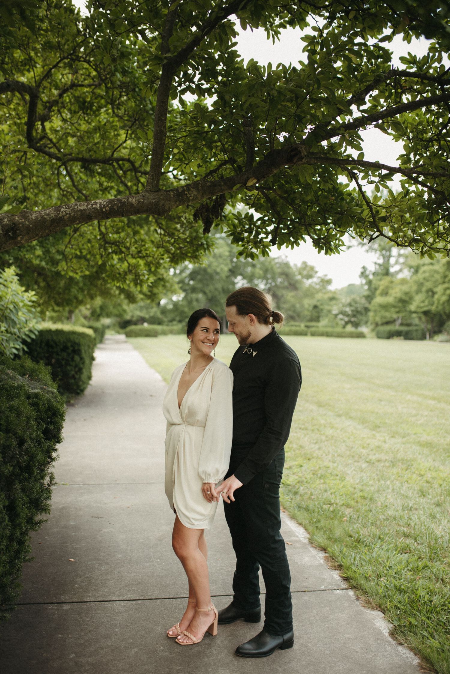 A couple stands beneath overhanging tree branches on a garden pathway.