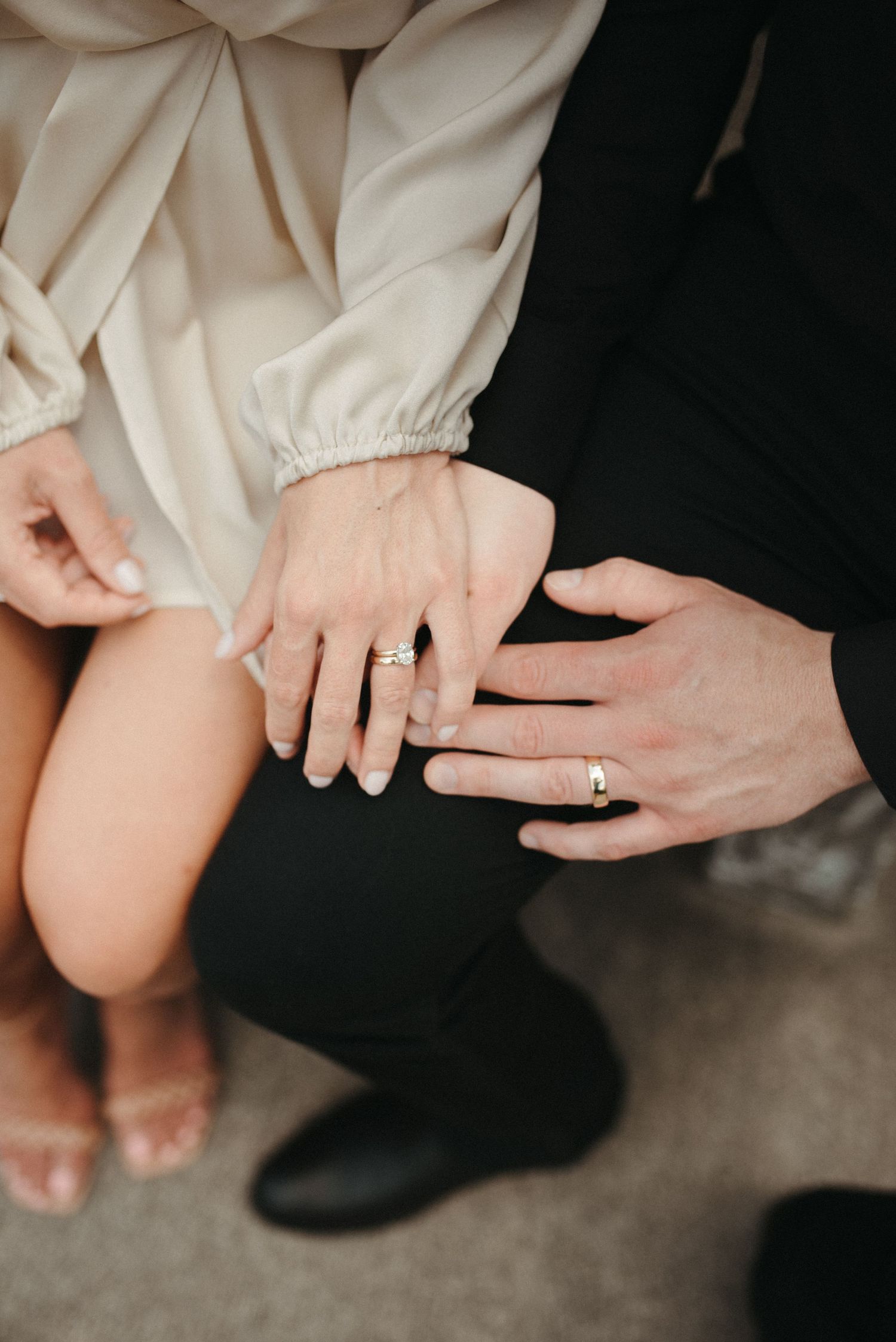 Close-up of hands with engagement rings shown against white dress and black suit.