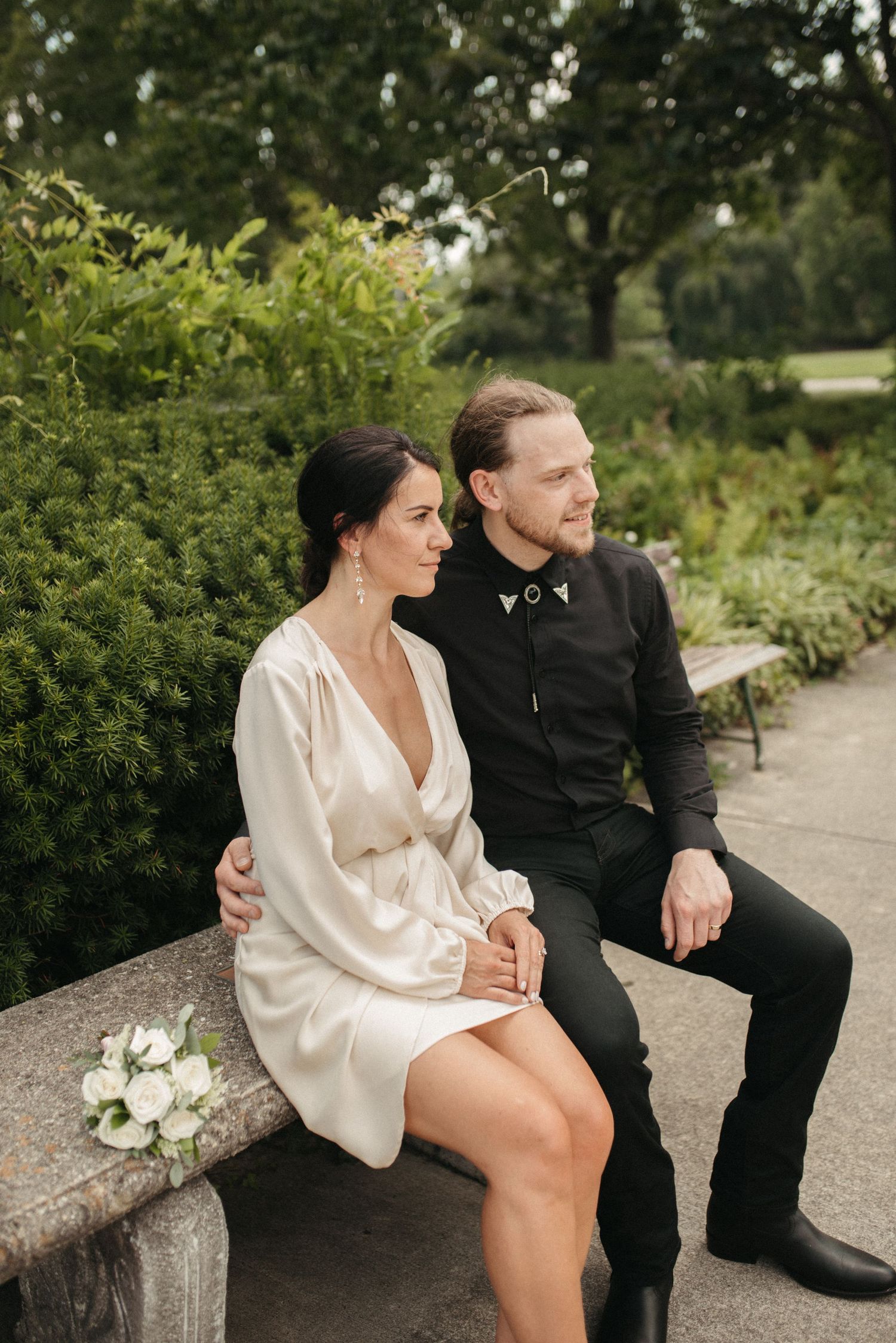 A couple sits on a stone bench in a garden wearing formal black and white attire.