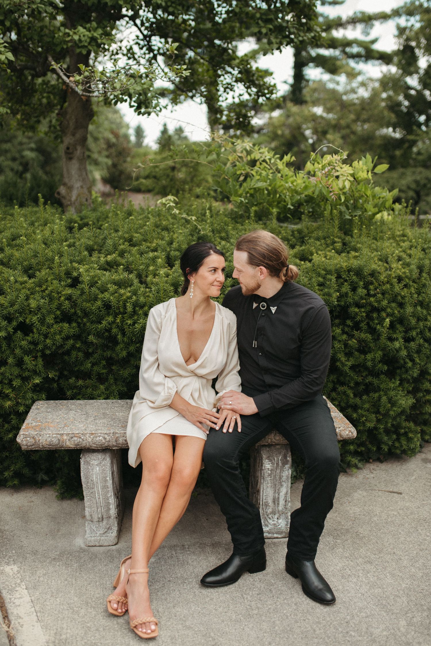 Couple in contrasting formal wear sits on stone bench surrounded by lush garden greenery.