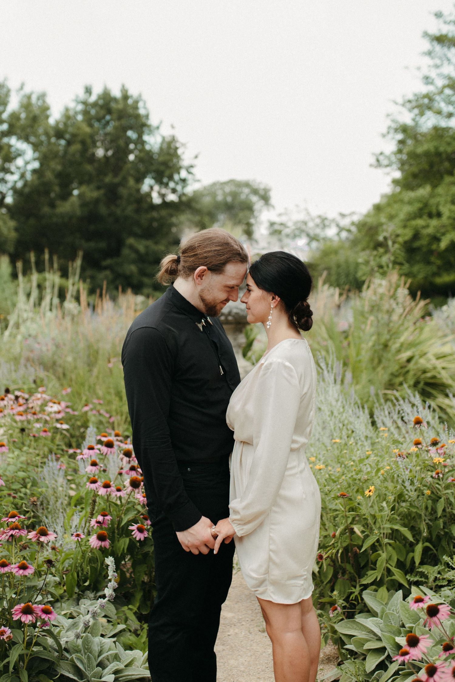 Romantic garden portrait of couple standing among pink coneflowers wearing formal attire.