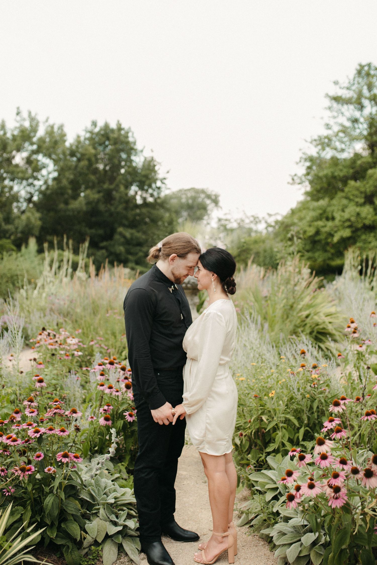 Intimate portrait of couple standing in flowering garden path wearing contrasting formal wear.