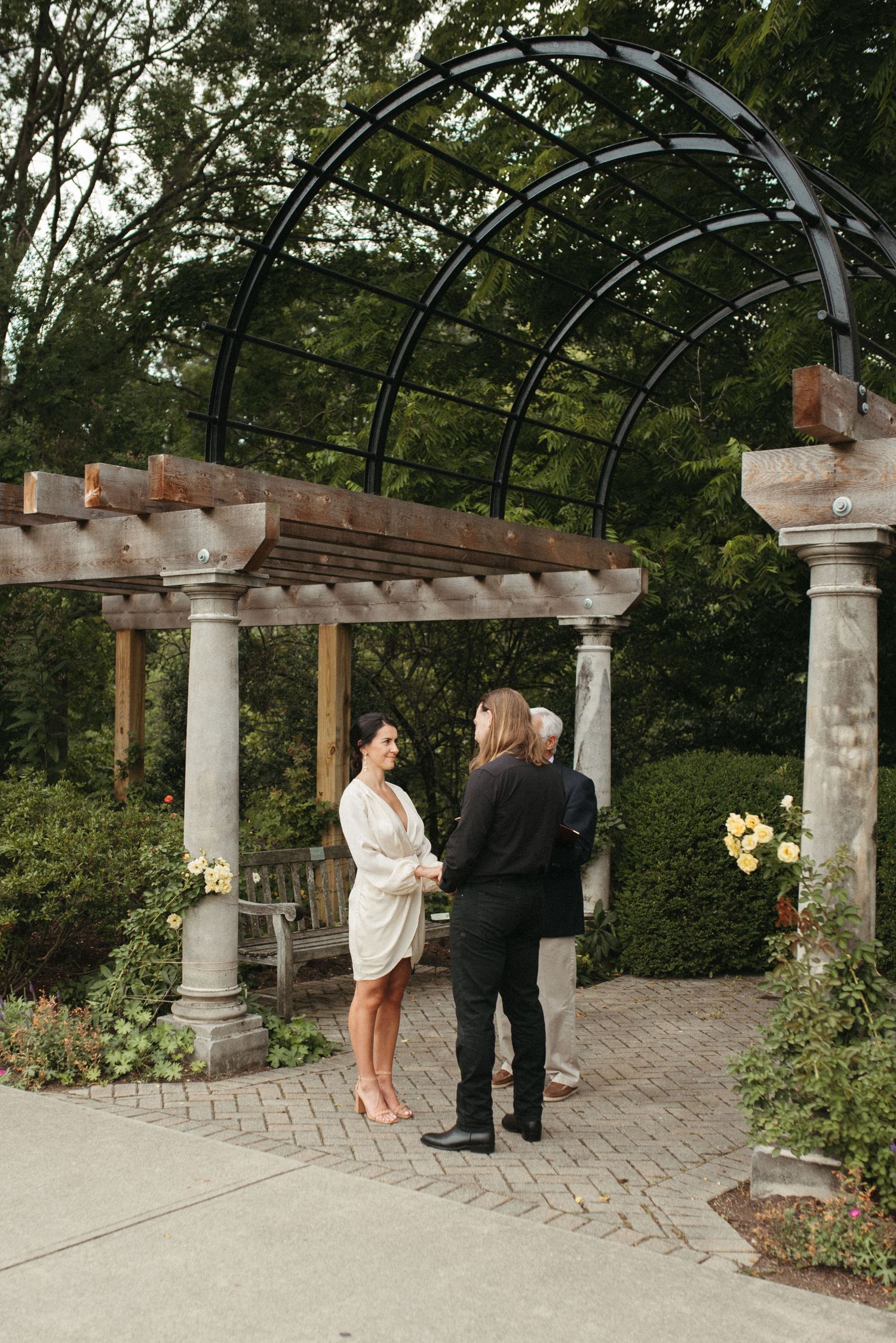 A wedding ceremony takes place under an arched garden structure with stone columns.