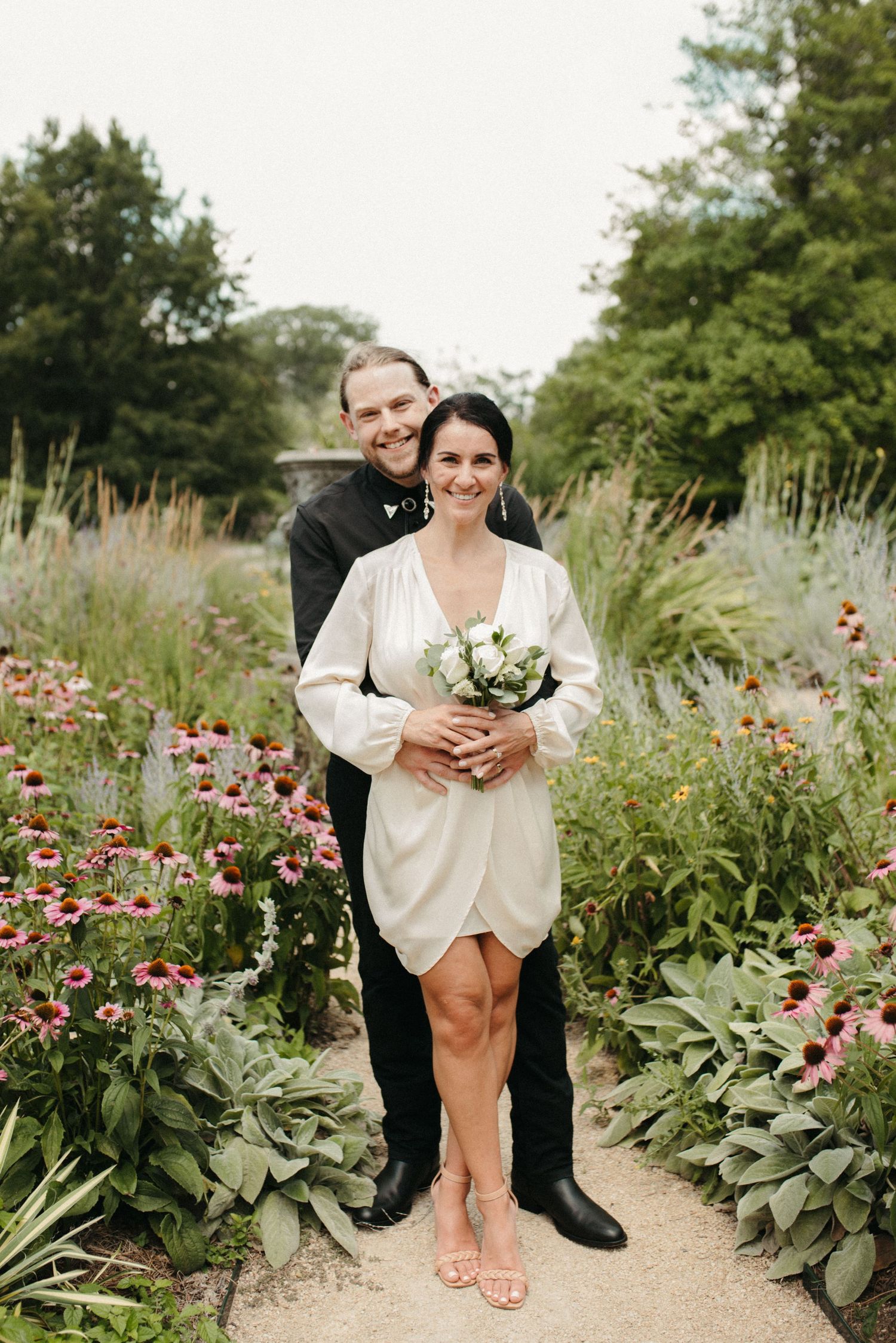 A couple poses in a garden path surrounded by pink flowers and greenery.