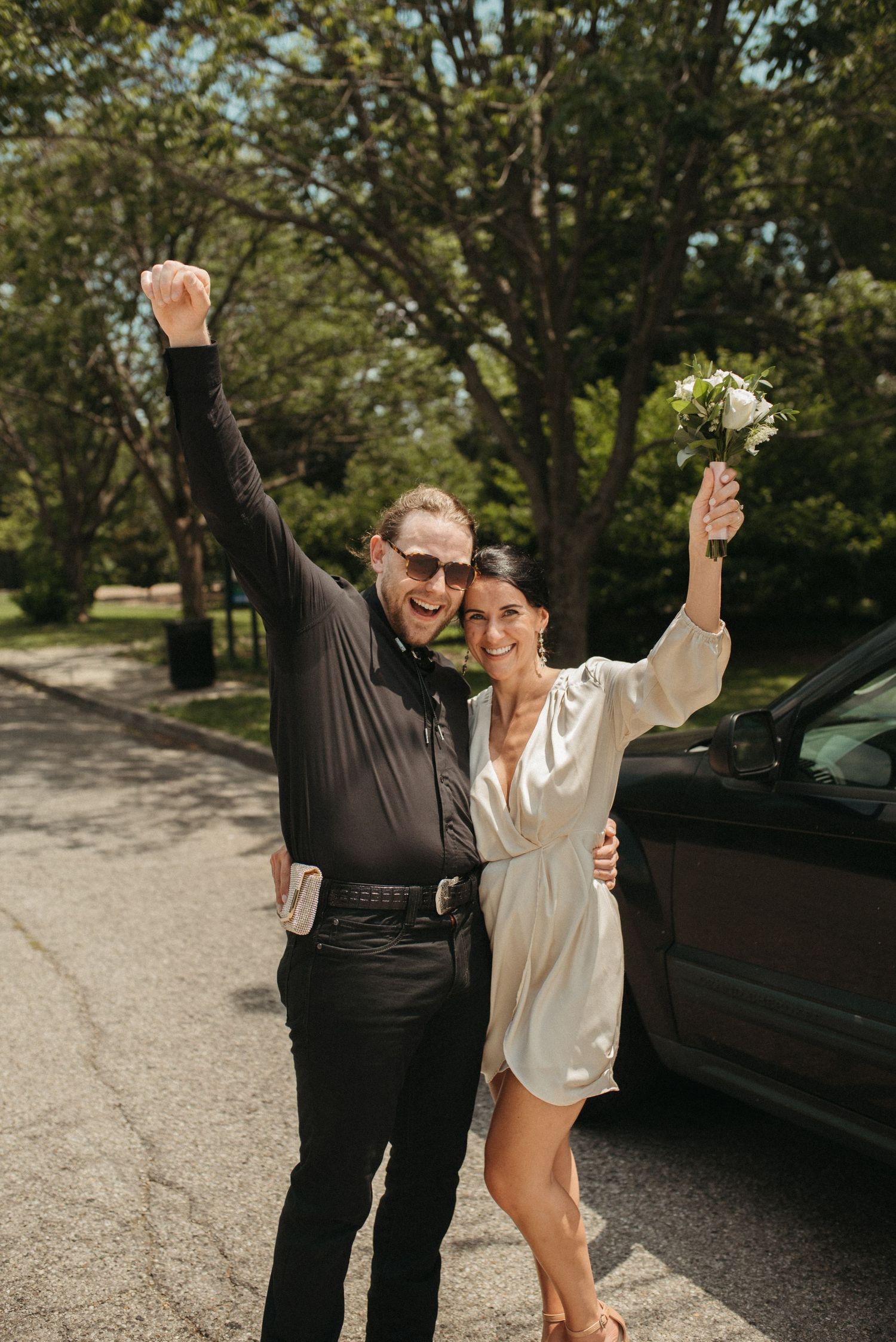 A celebratory couple raises their arms in joy on a tree-lined path.