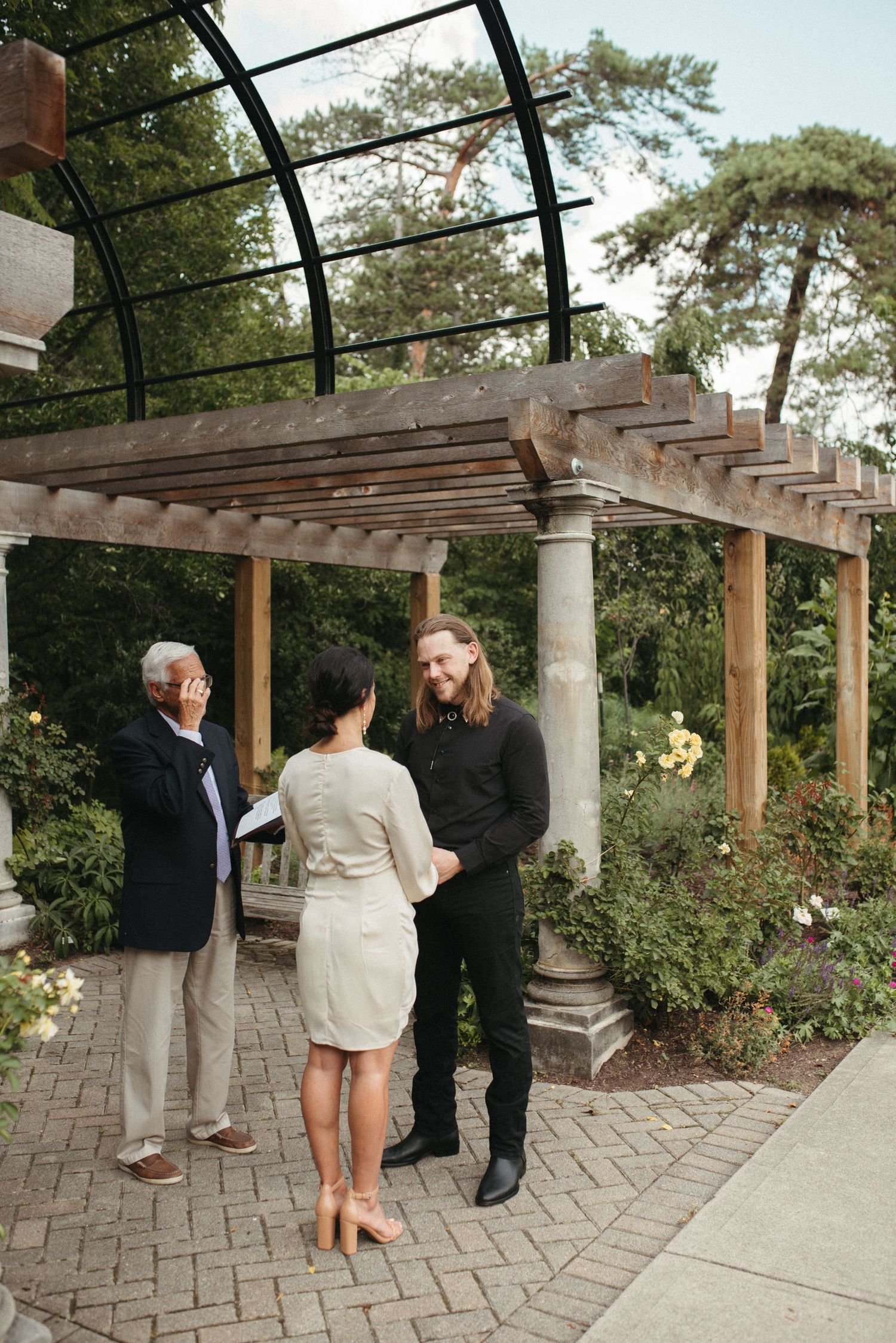 A wedding ceremony unfolds beneath a pergola with curved metal beams and stone pillars.