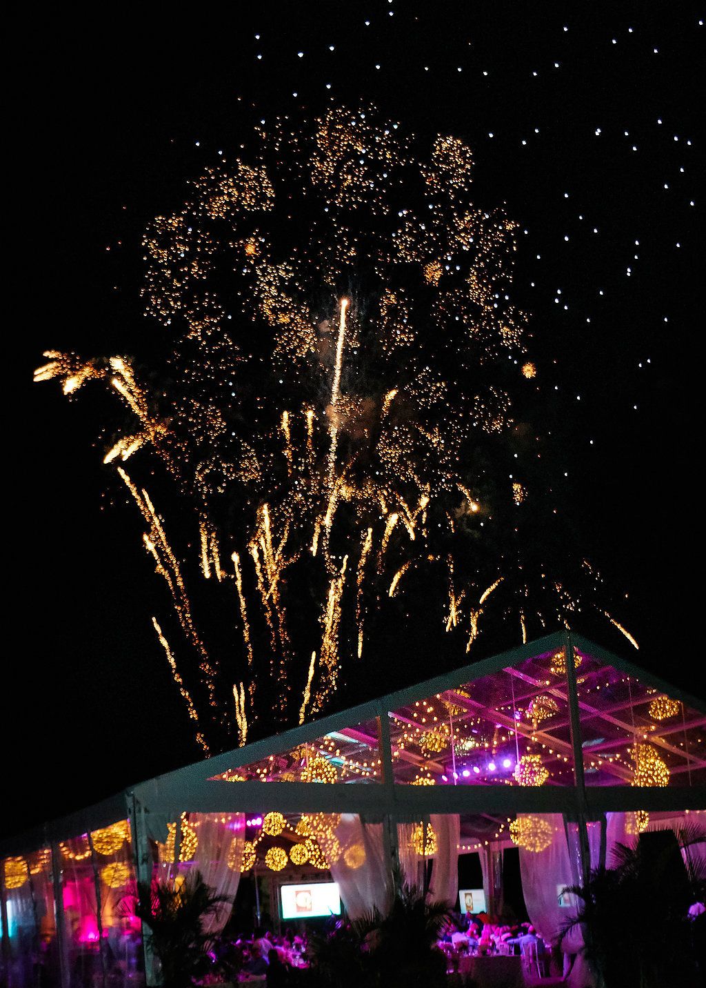 Fireworks display over an illuminated event tent with purple and gold lighting.