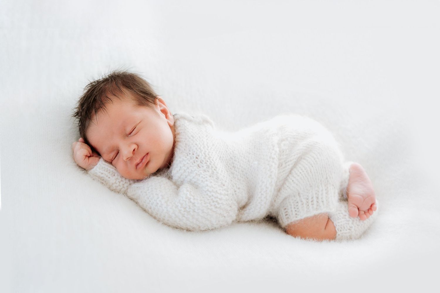 A sleeping newborn baby wearing a soft white sweater lies peacefully on a white background. In Redcliffe photographer and Mango hill studio