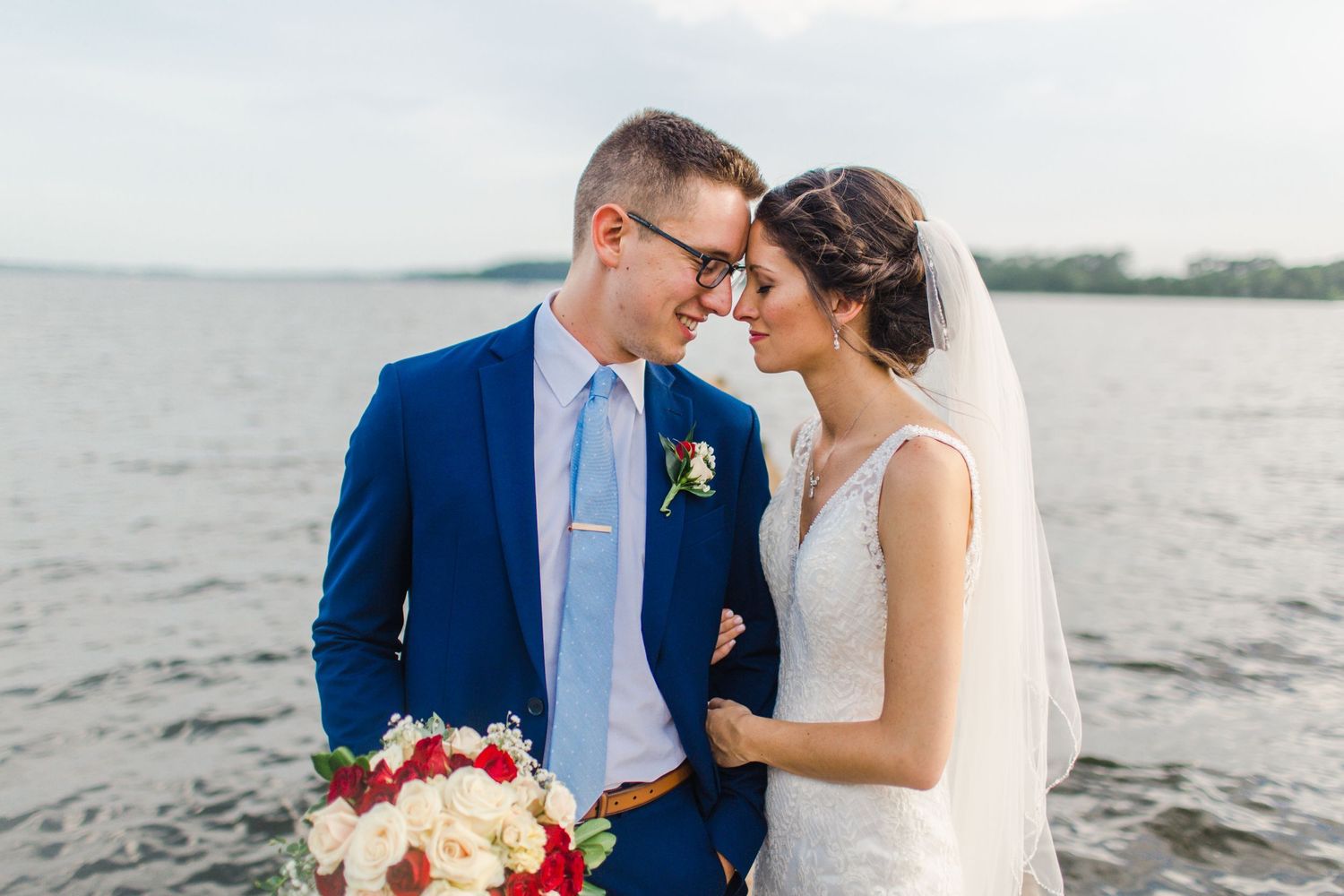 Waterfront wedding portrait with couple in blue suit and white dress holding red bouquet.