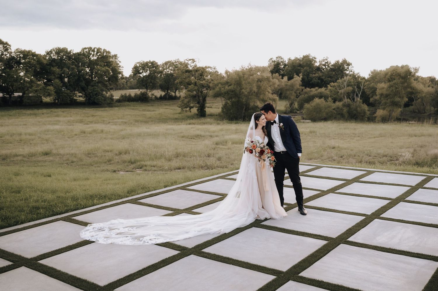 A couple stands together on a geometric tile patio surrounded by lush green fields during an outdoor wedding.