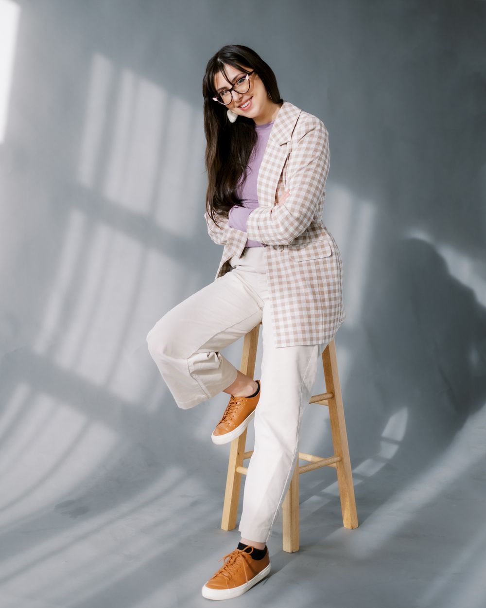 Relaxed lifestyle portrait on wooden stool featuring gingham jacket and white pants ensemble.