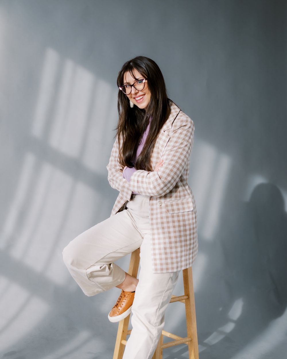 Casual studio portrait showcasing checkered jacket, white pants and tan sneakers with window light.
