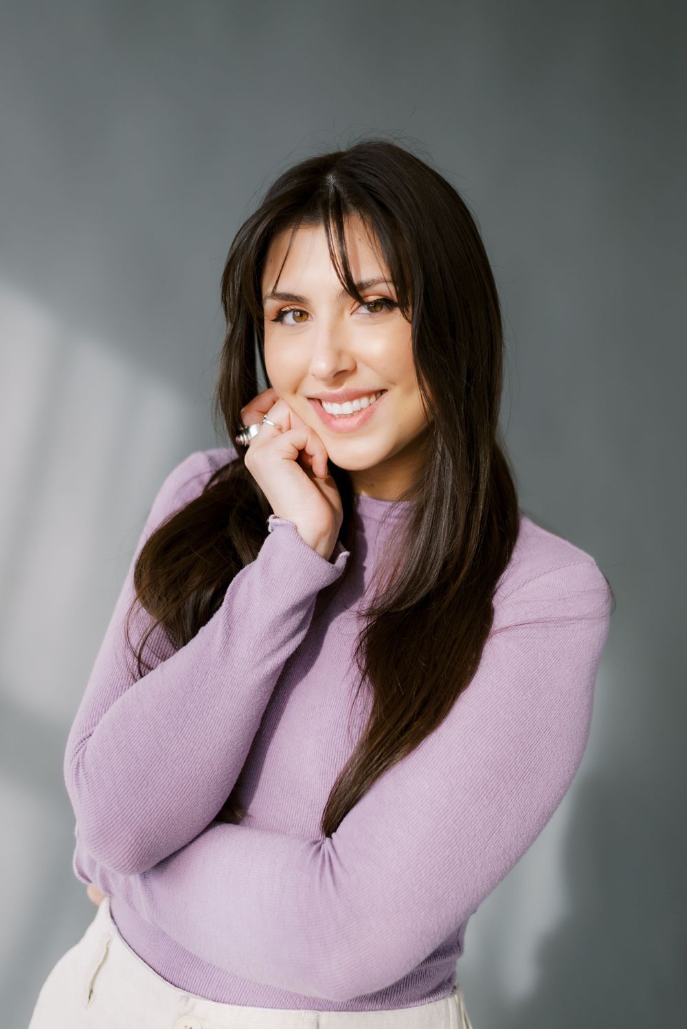 A portrait of someone wearing a lavender sweater and smiling warmly against a gray background.
