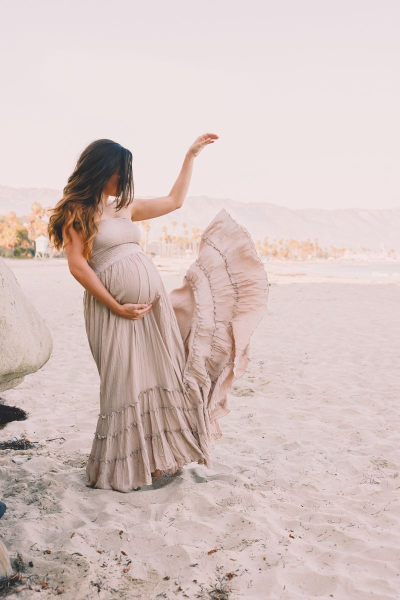 Boho maternity photo with flowing beige dress on sandy beach at sunset.