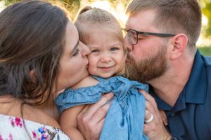 Family shares sweet kisses with toddler in blue dress during golden hour photo session.